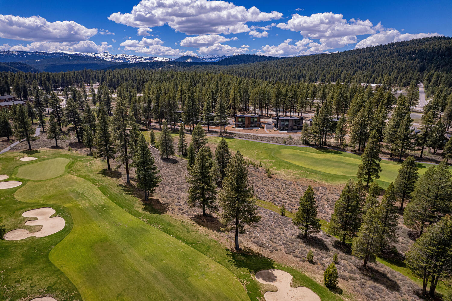 This aerial shot showcases a beautiful property nestled within a scenic landscape. The property features modern homes surrounded by lush greenery, including a golf course and dense forest. Snow-capped mountains can be seen in the distance, adding to the overall appeal of this tranquil setting.
