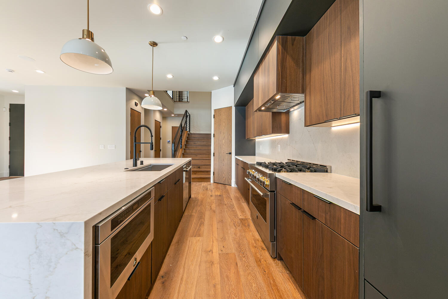 The image showcases a modern kitchen with wooden cabinetry and stainless steel appliances. A spacious island with a light countertop provides ample workspace, complemented by pendant lighting. The hardwood flooring adds warmth and elegance to the space, creating a sleek and inviting atmosphere.