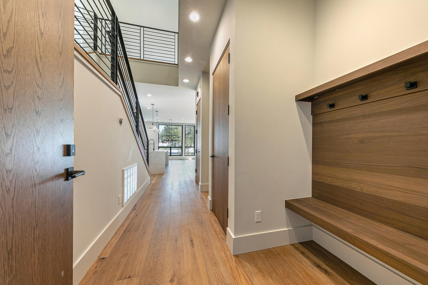 This interior shot showcases a modern home's hallway and staircase. The hardwood flooring leads the eye through the space, complemented by a minimalist staircase with black railings. A built-in bench and coat rack are present on the right, enhancing functionality and design.