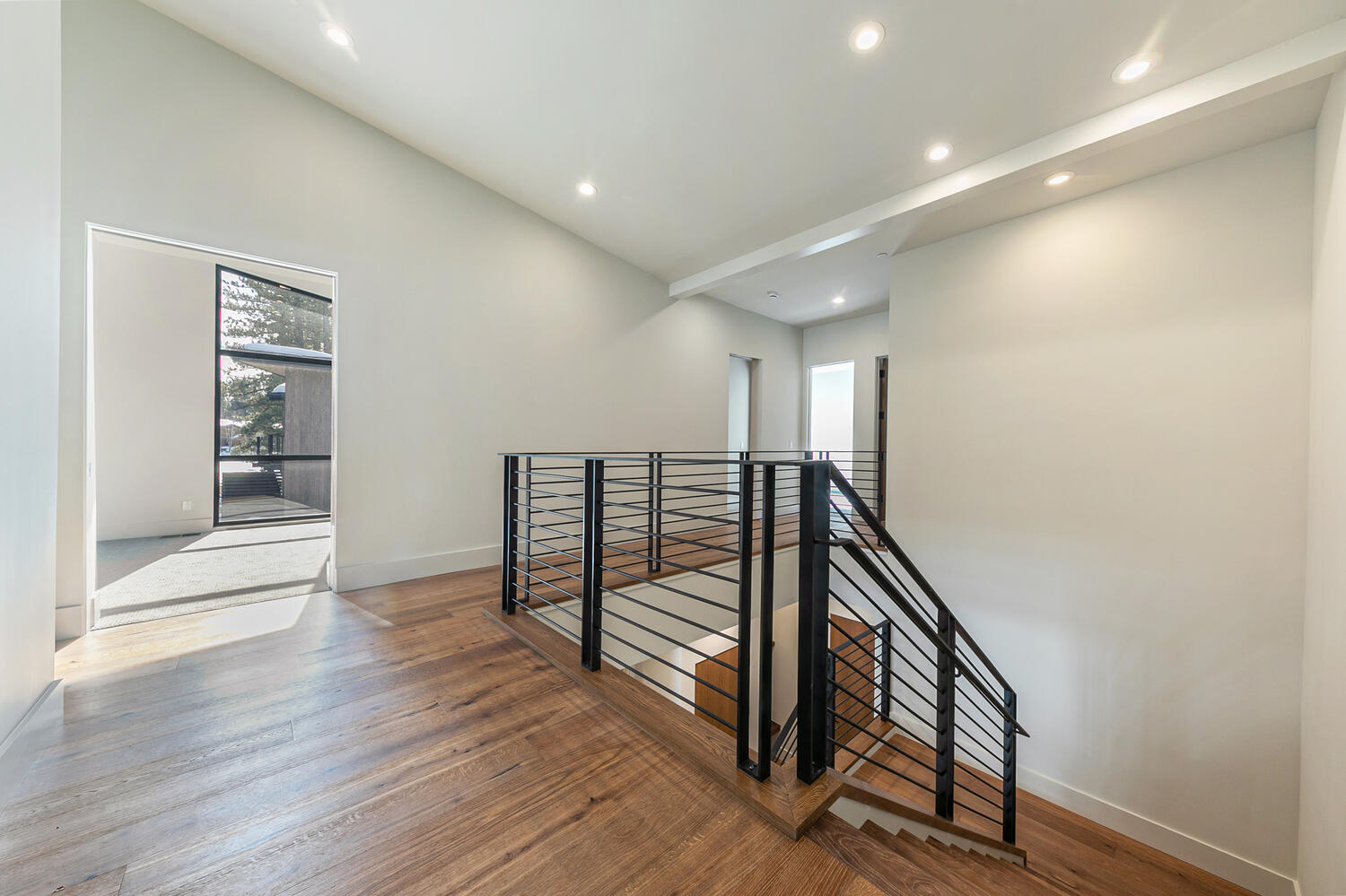 This is an interior shot of a hallway and stairs in a modern home. The hallway features hardwood flooring and leads to a light-filled room visible through a doorway. The staircase has a contemporary black metal railing and wood steps, contributing to the home's stylish aesthetic.