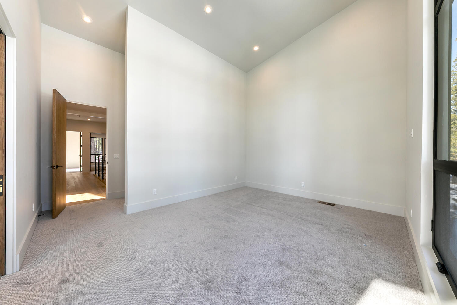 This is a spacious primary bedroom featuring neutral-toned carpet and light gray walls. Recessed lighting brightens the room, and a large doorway provides access to another area. A large window allows for natural light to enter the space.