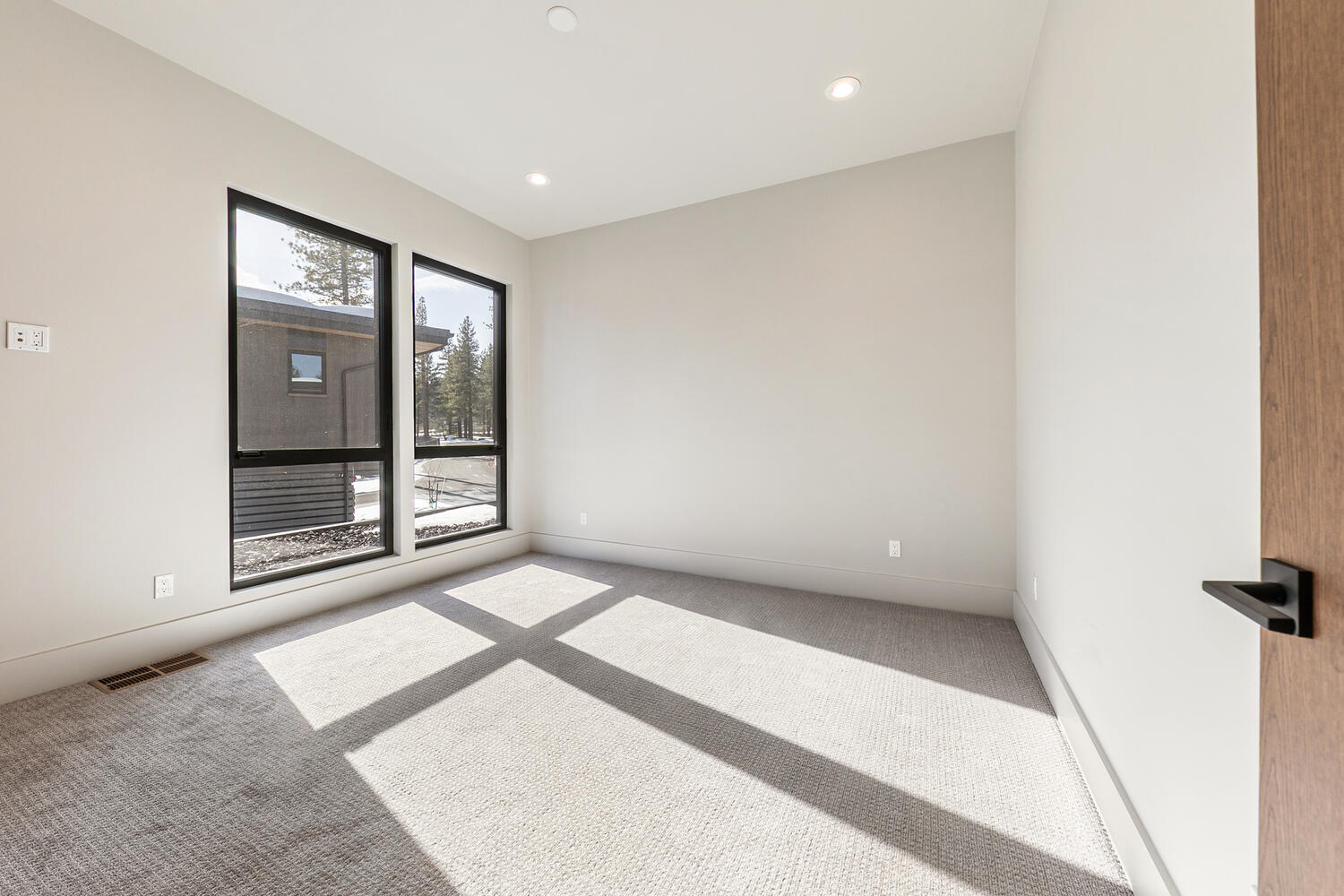 This is a shot of a bright, modern bedroom with gray walls and carpet. Two large windows provide natural light and views of the surrounding landscape. A partial view of the door gives a peek at the wood finish and hardware.