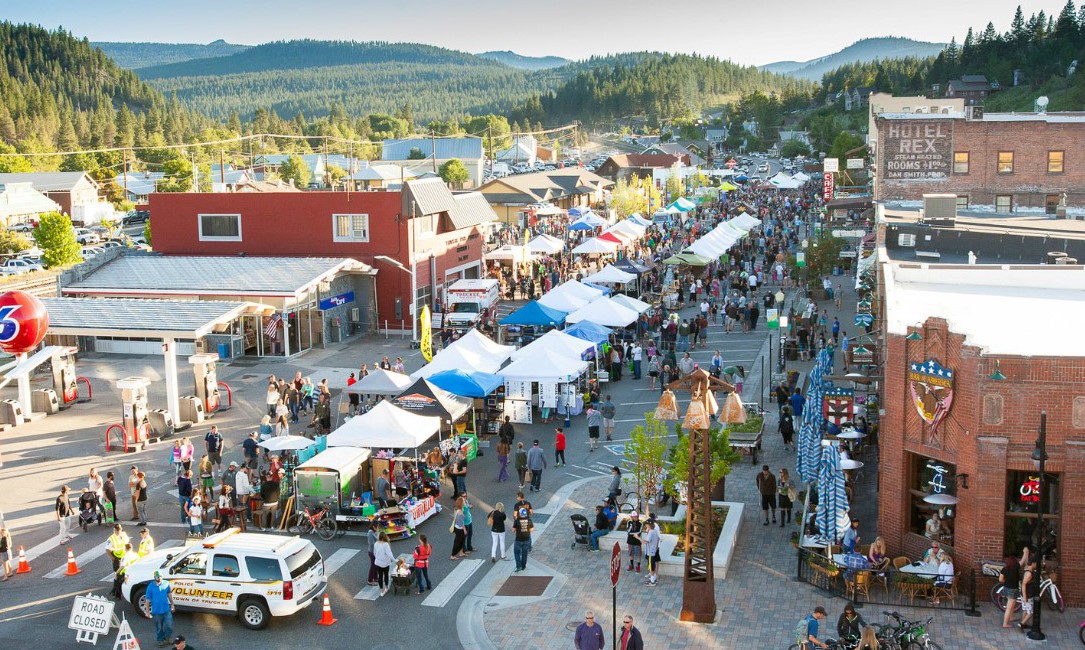 This is an aerial view of a vibrant street fair in a small town. The scene is bustling with people, vendor tents, and local businesses. The image captures a lively community atmosphere, showcasing potential for high foot traffic and local charm.