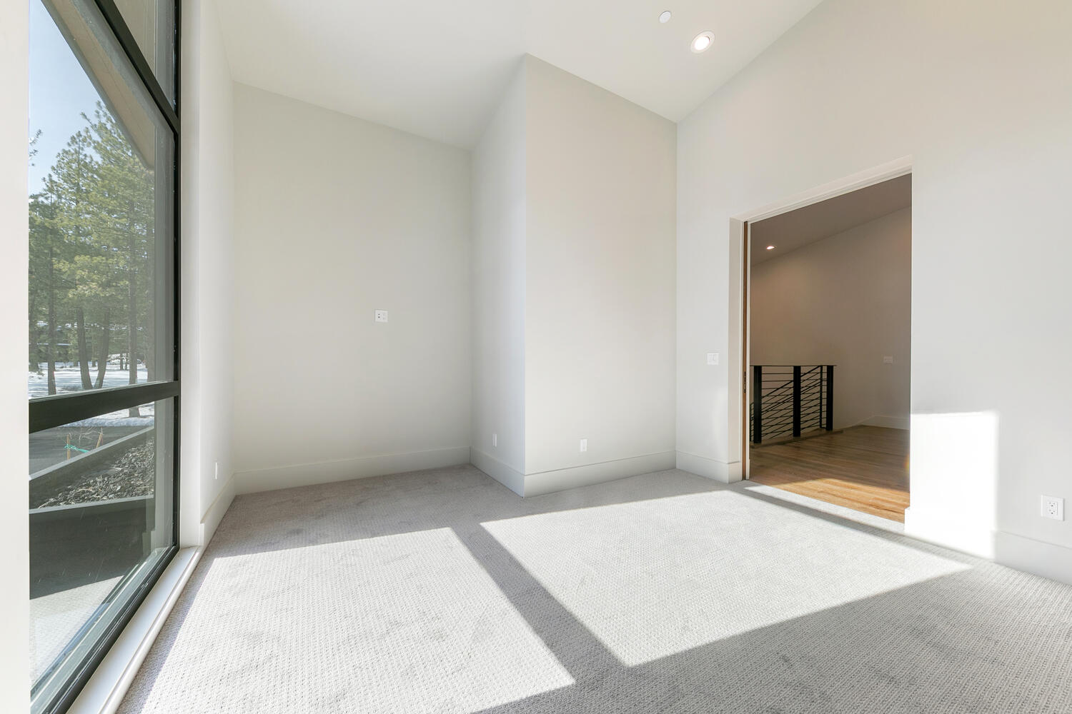 This is an interior shot of a bright, empty bedroom featuring neutral carpet and light gray walls. A large window to the left provides natural light and a view of trees outdoors. An open doorway to the right provides a glimpse of another room and a staircase.