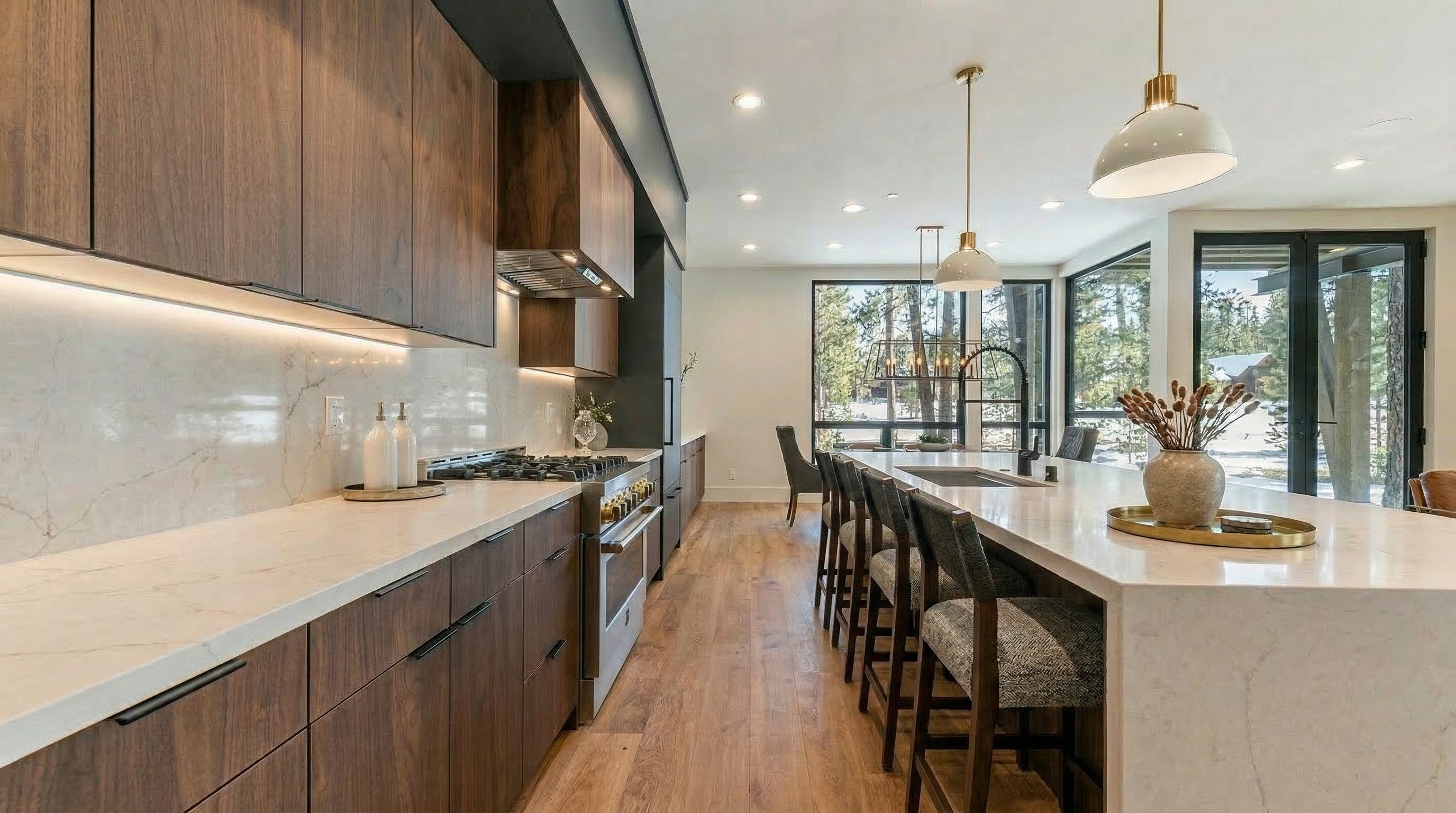 This is a modern kitchen featuring dark wood cabinets and light countertops with a waterfall island. Pendant lights hang above the island. Large windows provide ample natural light and views to the trees outside.