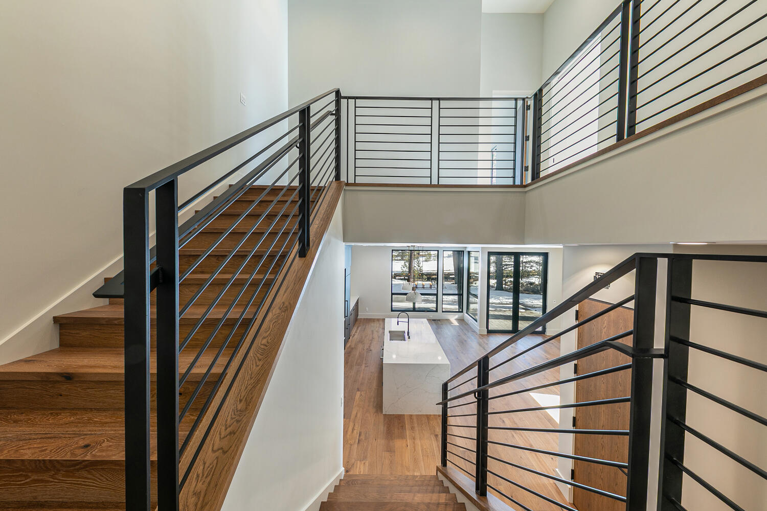 The image showcases a well-lit, modern interior with a striking staircase. The wooden stairs contrast with the black metal railings, creating a contemporary design. Below, a glimpse of the kitchen with an island can be seen through nearby windows.
