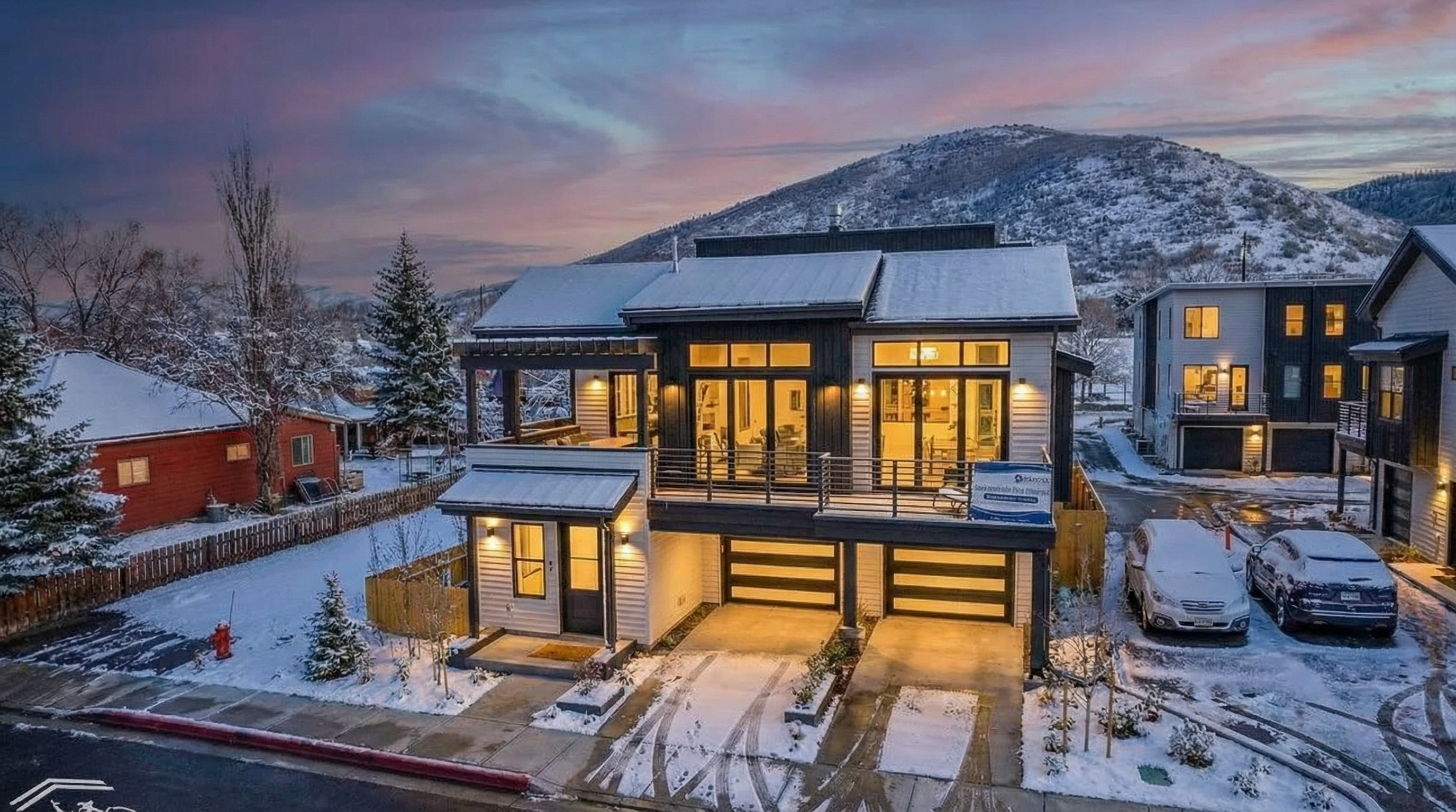 This image showcases the front exterior of a modern, two-story home in a winter setting. The architecture features a combination of dark and light siding, large windows, and a balcony. Double garage doors are visible at the ground level, and the property is surrounded by a snow-covered landscape, giving it a serene and luxurious appeal.