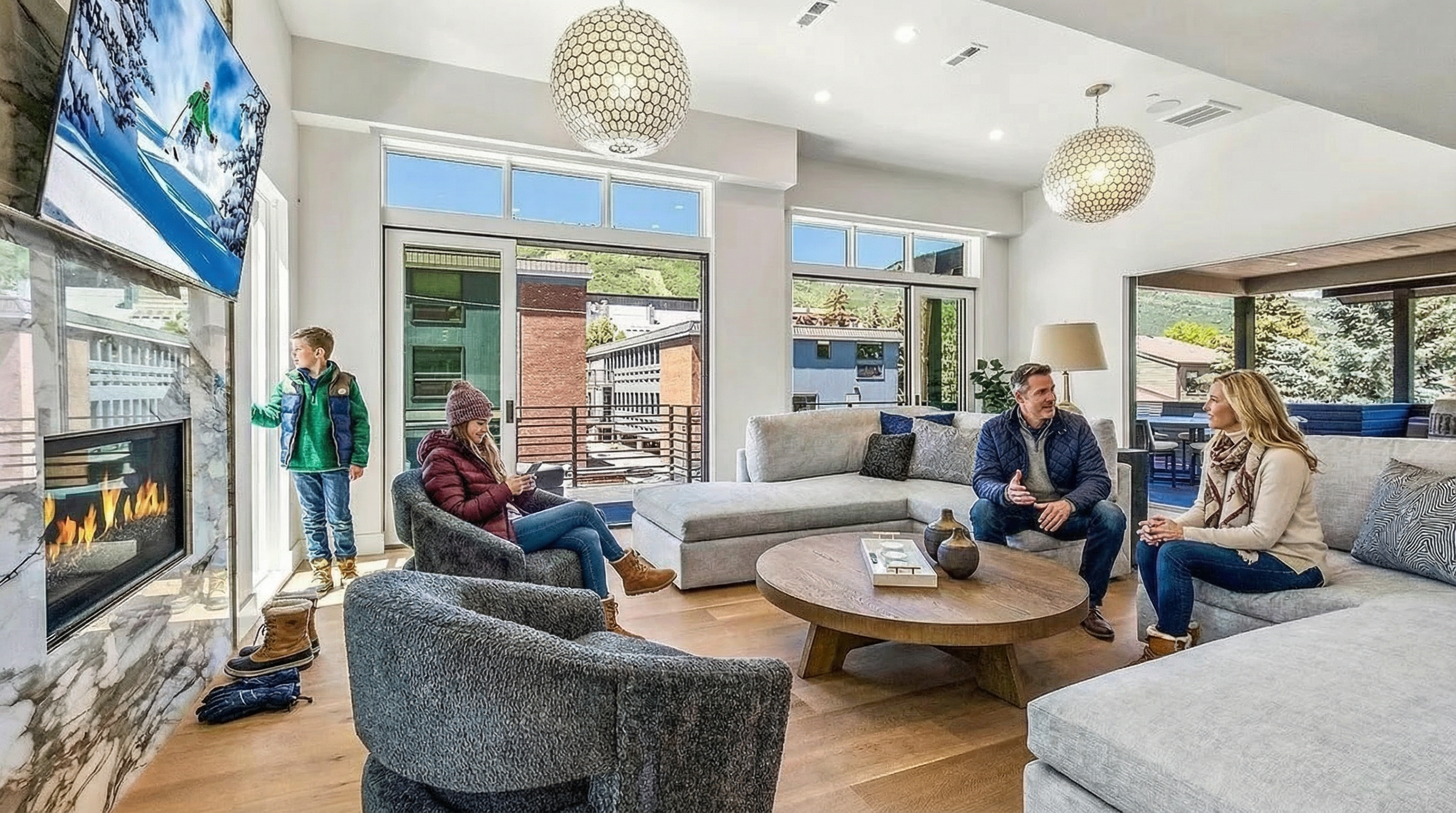 This is a well-lit living room, showcasing a comfortable and inviting atmosphere. It features a modern fireplace, comfortable seating arrangements with a sectional sofa and plush chairs, and large windows that provide natural light and outdoor views. The space is decorated with contemporary light fixtures and a wooden coffee table, contributing to a sophisticated yet relaxed living environment.