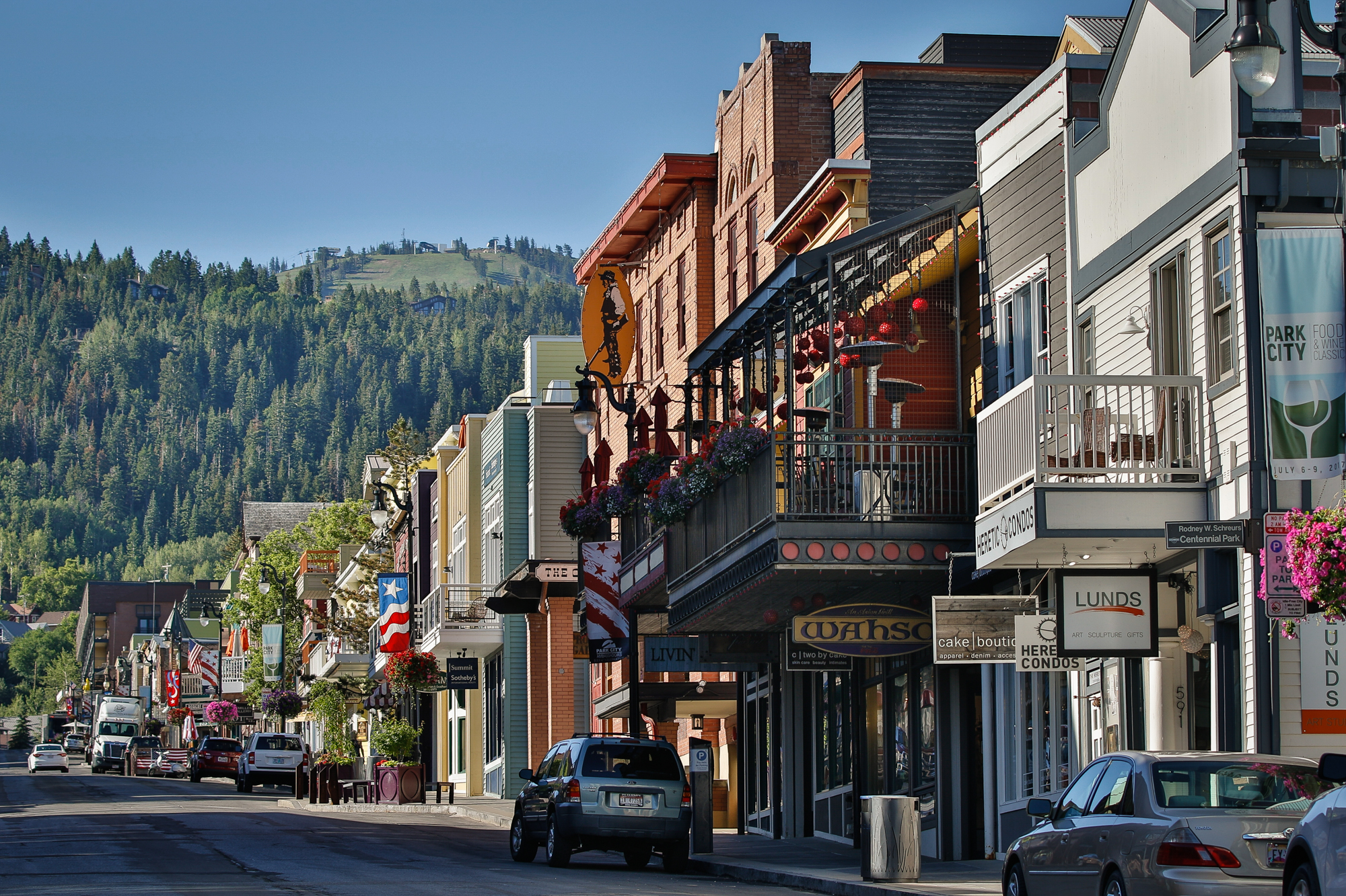 This image showcases a charming street view with commercial buildings exhibiting unique architectural details and inviting storefronts. Balconies adorned with flowers add a touch of elegance, while parked cars suggest a bustling city center. The backdrop of tree-covered hills adds a scenic context, enhancing the appeal of this desirable location.