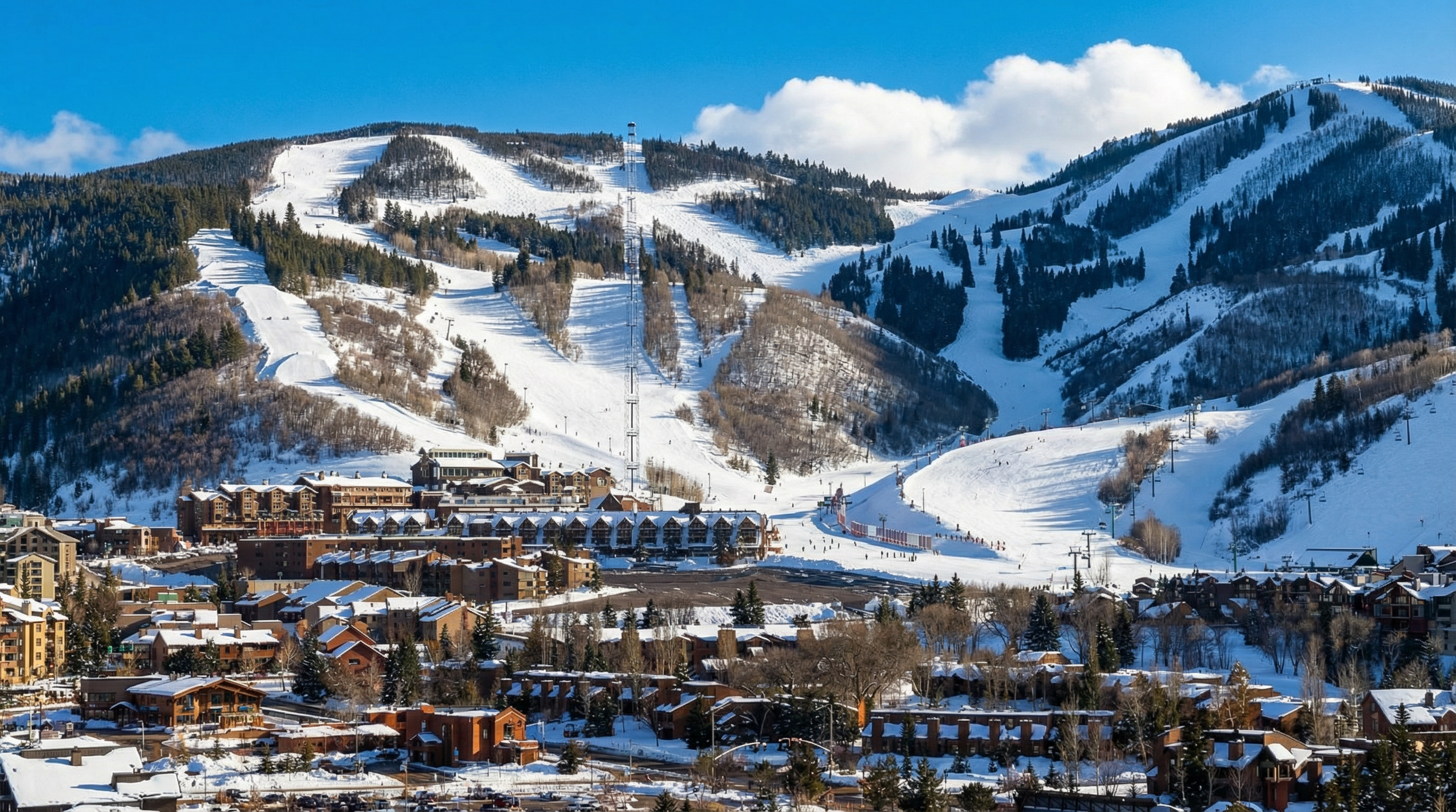 This aerial shot captures a ski resort town nestled at the base of snow-covered mountains. The foreground features various residential and commercial buildings, many with snow-covered roofs, surrounded by trees. Ski slopes are visible on the mountain, creating a picturesque and desirable location for winter sports enthusiasts looking for real estate opportunities.