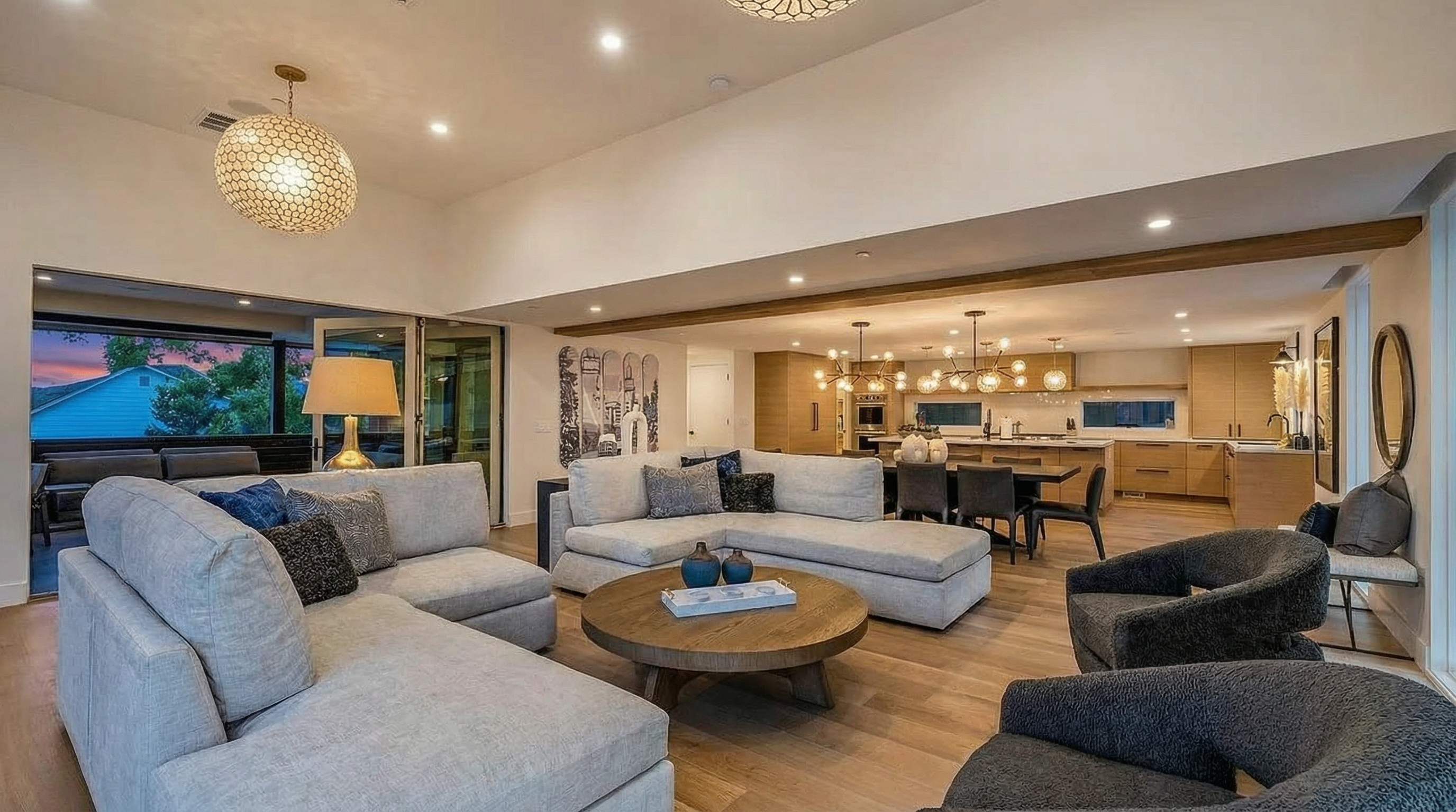 This is an interior shot of a living room that seamlessly connects to a kitchen. The living room features a large, light-colored sectional sofa, round wooden coffee table, and comfortable armchairs. The open floor plan is enhanced by wooden flooring and ample natural light creating a welcoming and modern atmosphere suitable for entertaining or relaxing.