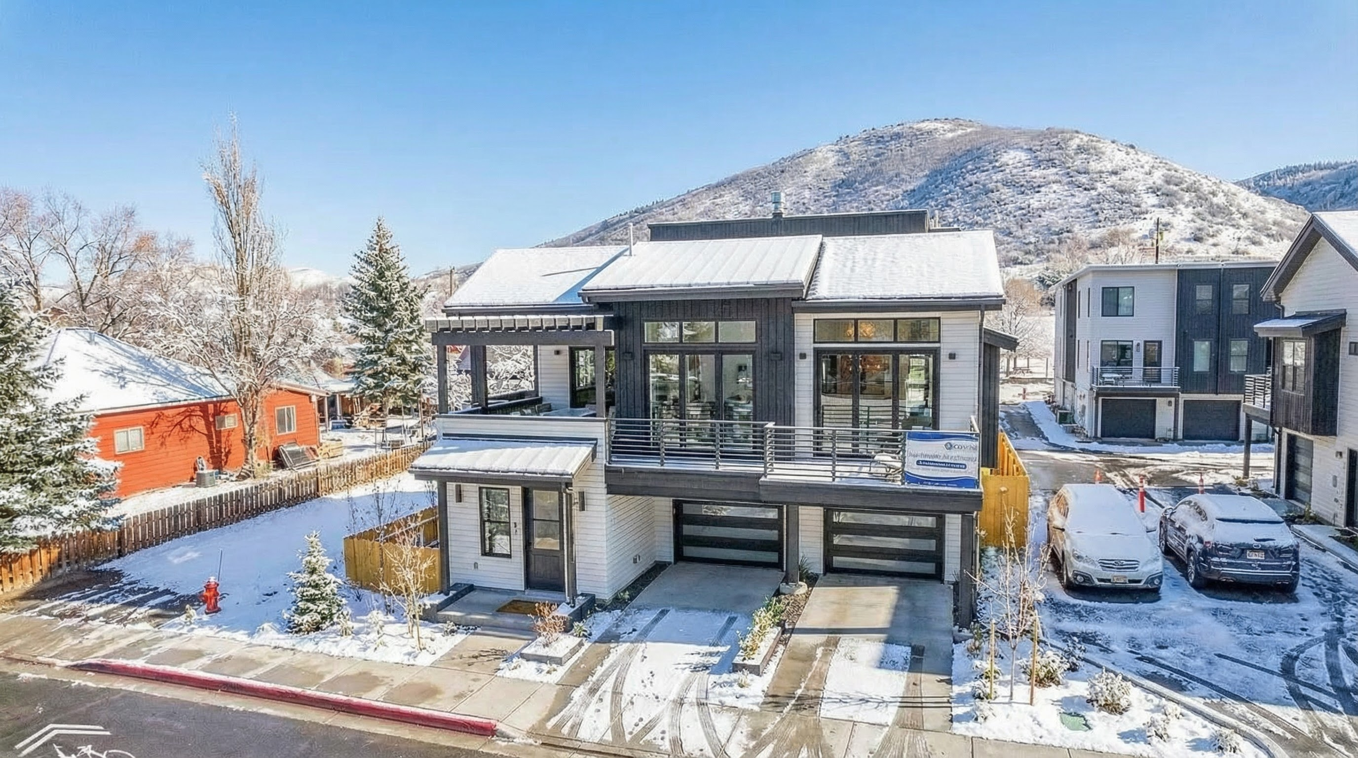 This is a front view of a modern two-story home in a snowy setting. It features a combination of white and dark gray siding, large windows, and a balcony. The house has a two-car garage and a landscaped front yard, presenting a contemporary and inviting curb appeal.