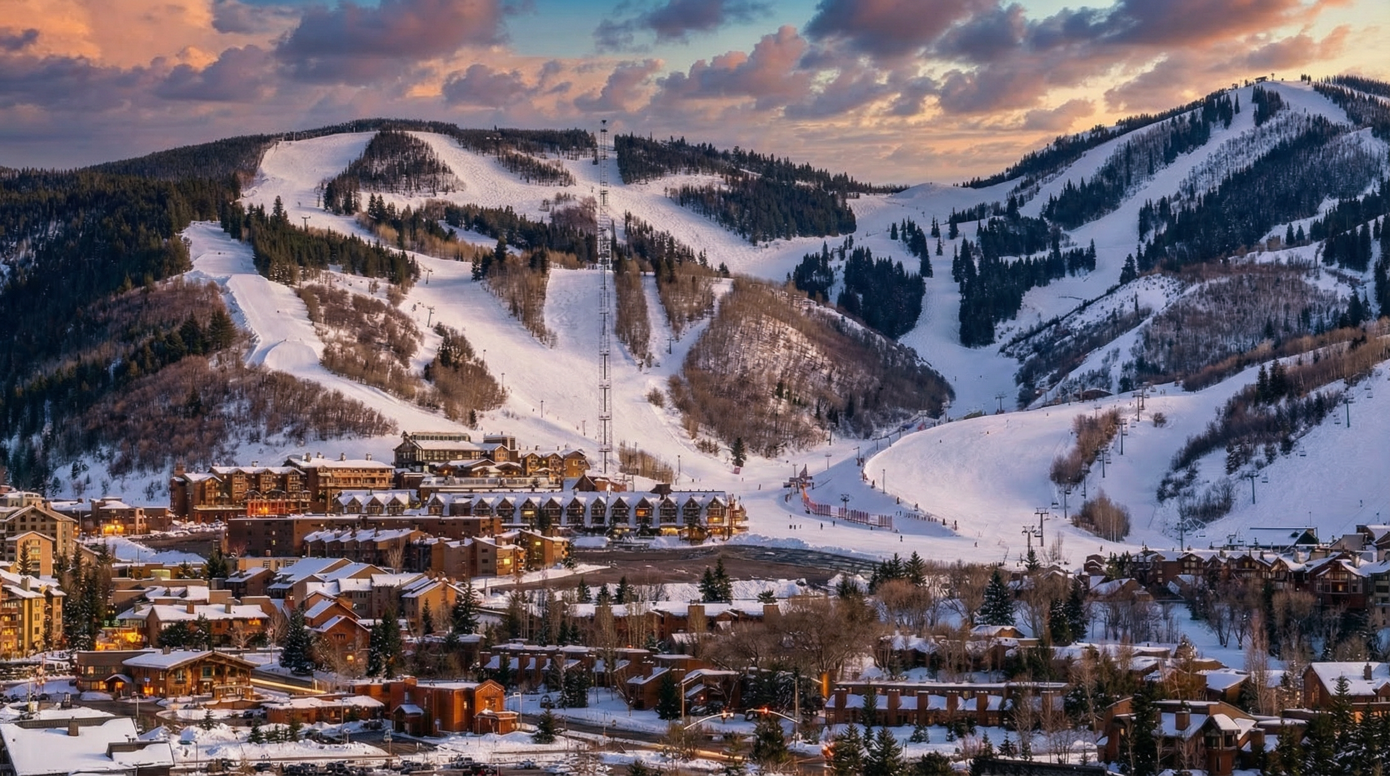 This image showcases a ski resort town nestled at the base of snow-covered mountains, likely depicting a desirable winter vacation destination. The scene includes residential buildings, a ski slope interspersed with evergreen trees, and a dramatic sky at dusk or dawn. The overall impression is one of luxury, recreation, and scenic beauty, making it ideal for promoting real estate in the area.