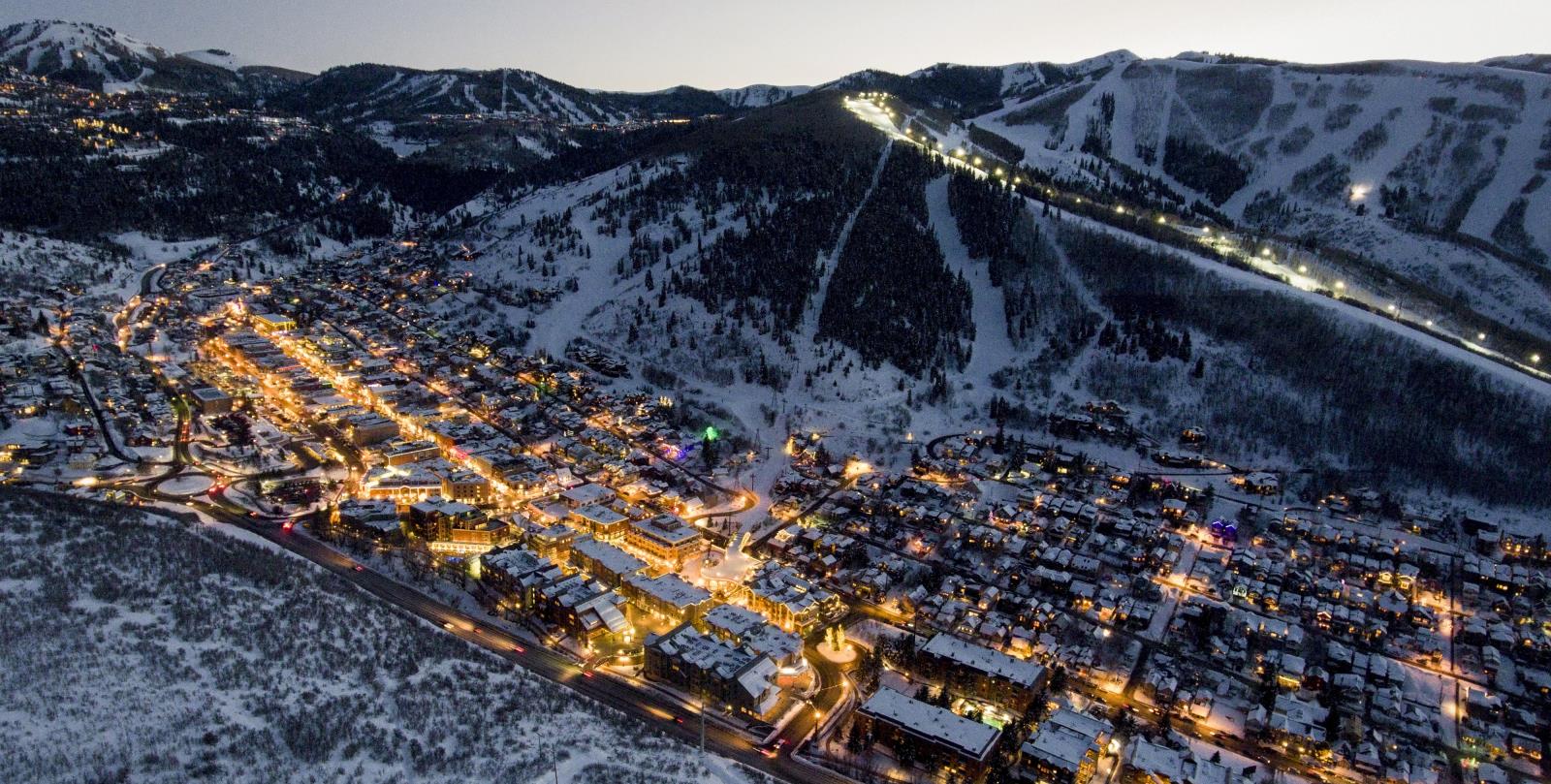 This aerial view showcases a snow-covered town nestled against a mountain backdrop. Lights from buildings and streetlights illuminate the scene creating a warm and inviting atmosphere. The image suggests a luxurious, ski-town lifestyle and highlights the desirable location amid natural beauty.