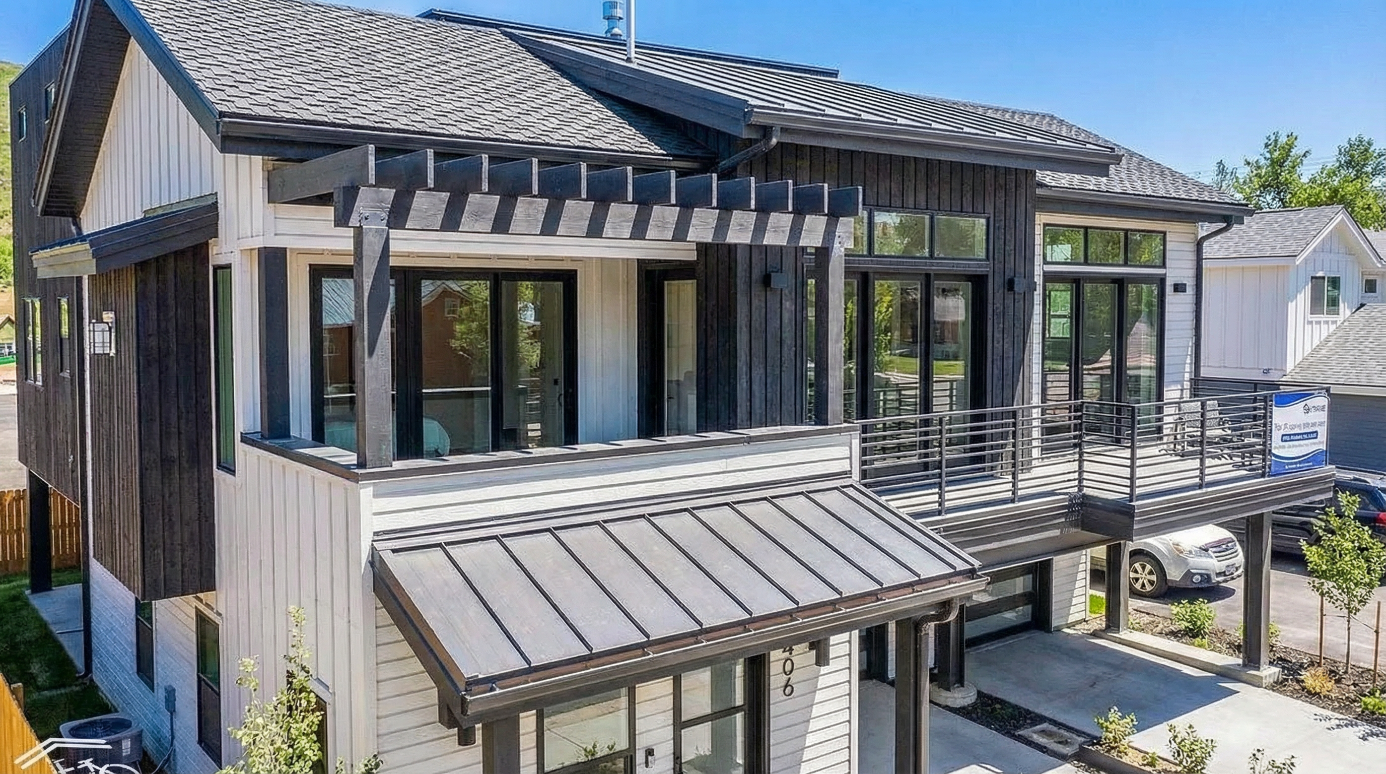 This is a front exterior view of a two-story, modern home with a striking architectural design. The house features a mix of white and dark wood siding, complemented by black-framed windows and a pergola detail over a balcony. The metal roof adds to the contemporary aesthetic, while a visible garage and well-maintained yard contribute to the property's overall appeal.