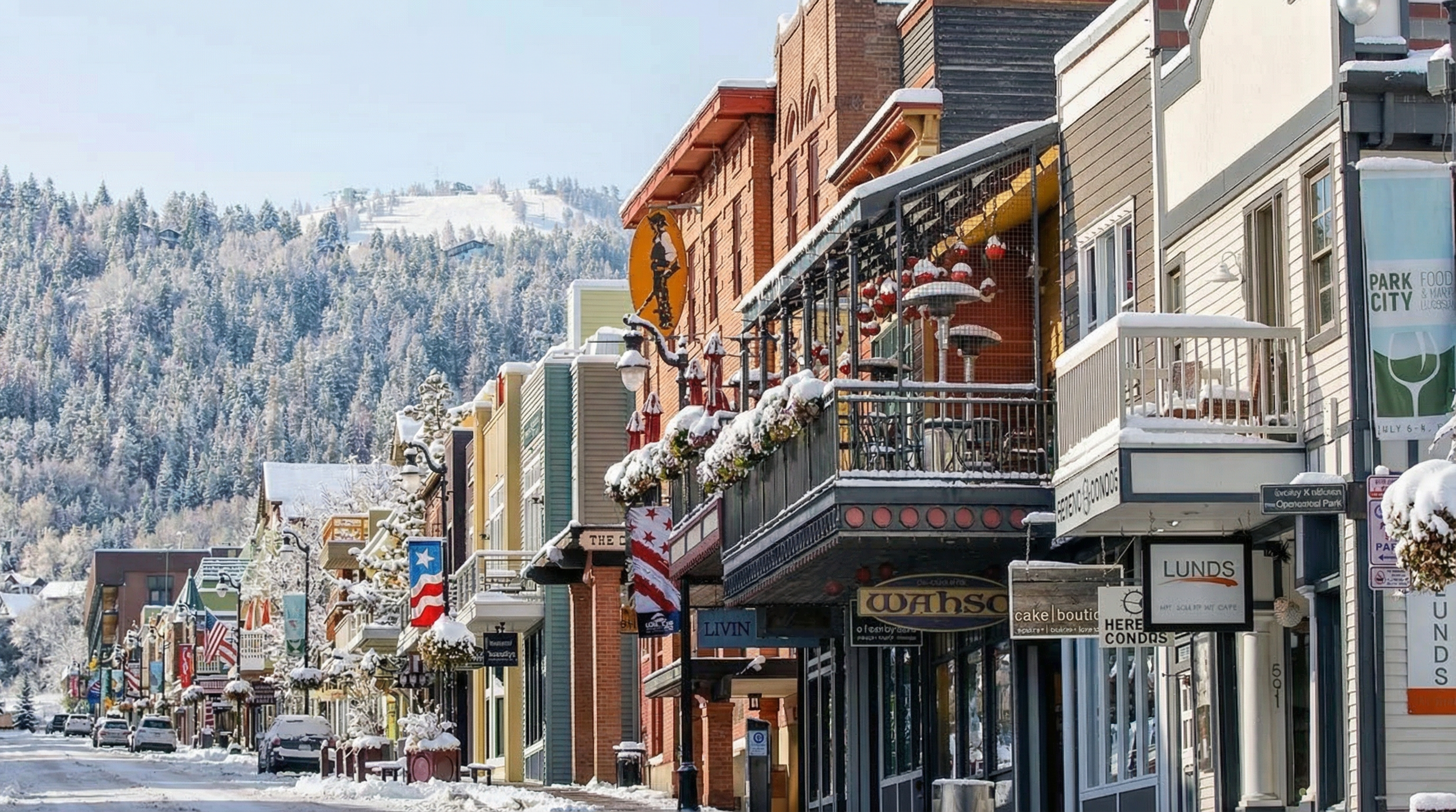 The image showcases a charming street in Park City during winter, lined with commercial buildings featuring a mix of brick and siding facades. Snow blankets the sidewalks and rooftops, lending a picturesque quality. Various storefront signs, including 'Wahso' and 'LUNDS', are visible, indicating a vibrant commercial district. The buildings exhibit architectural details like balconies and awnings.