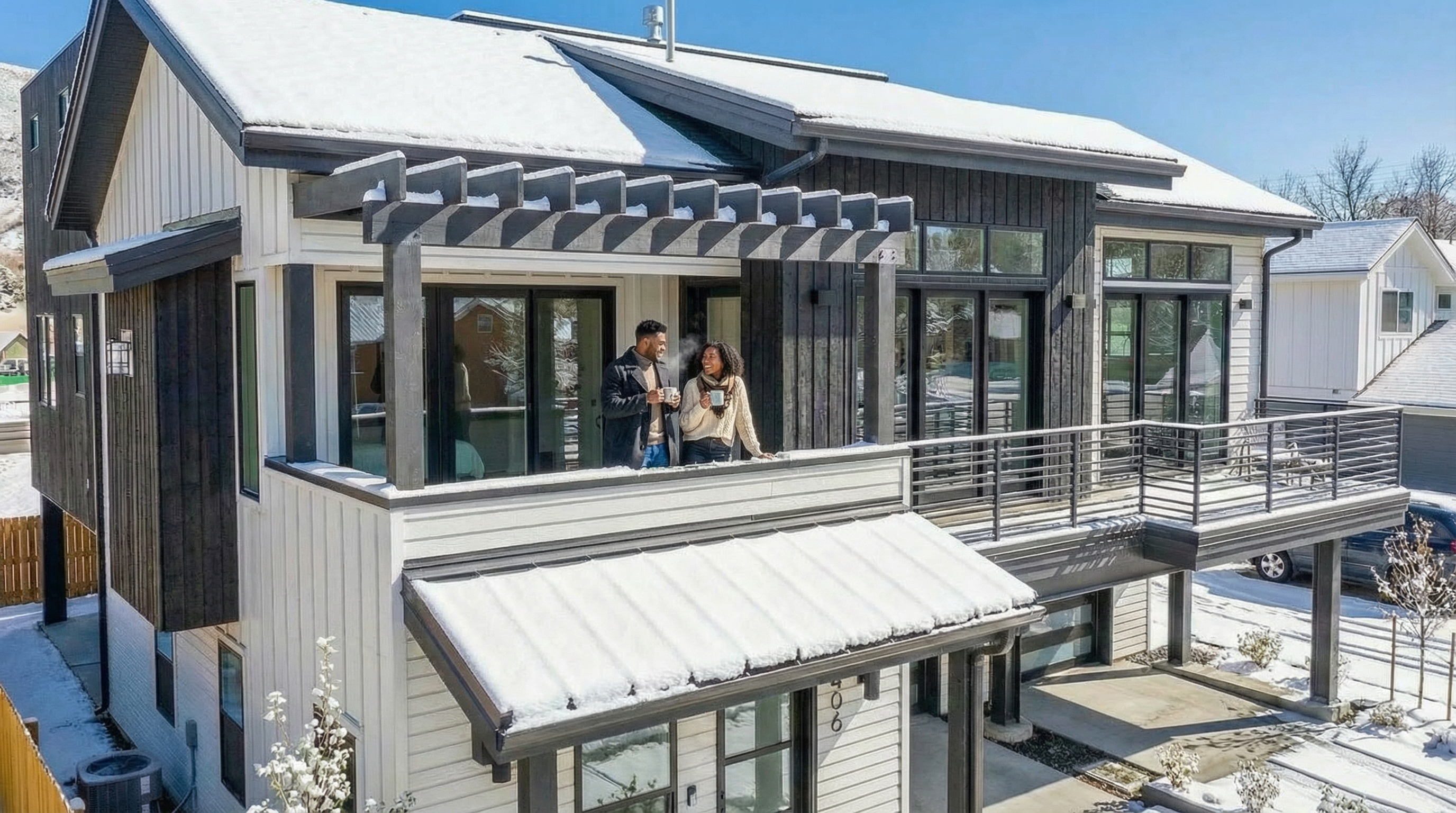 This is a front exterior view of a modern two-story home with a mixed material facade of white siding and dark wood paneling. The house features a balcony with a grey pergola above and a metal railing, and a covered entryway partially covered with snow. The residence is situated in a snowy landscape, adding to its cozy yet contemporary appeal, and offers a glimpse of an attached garage.