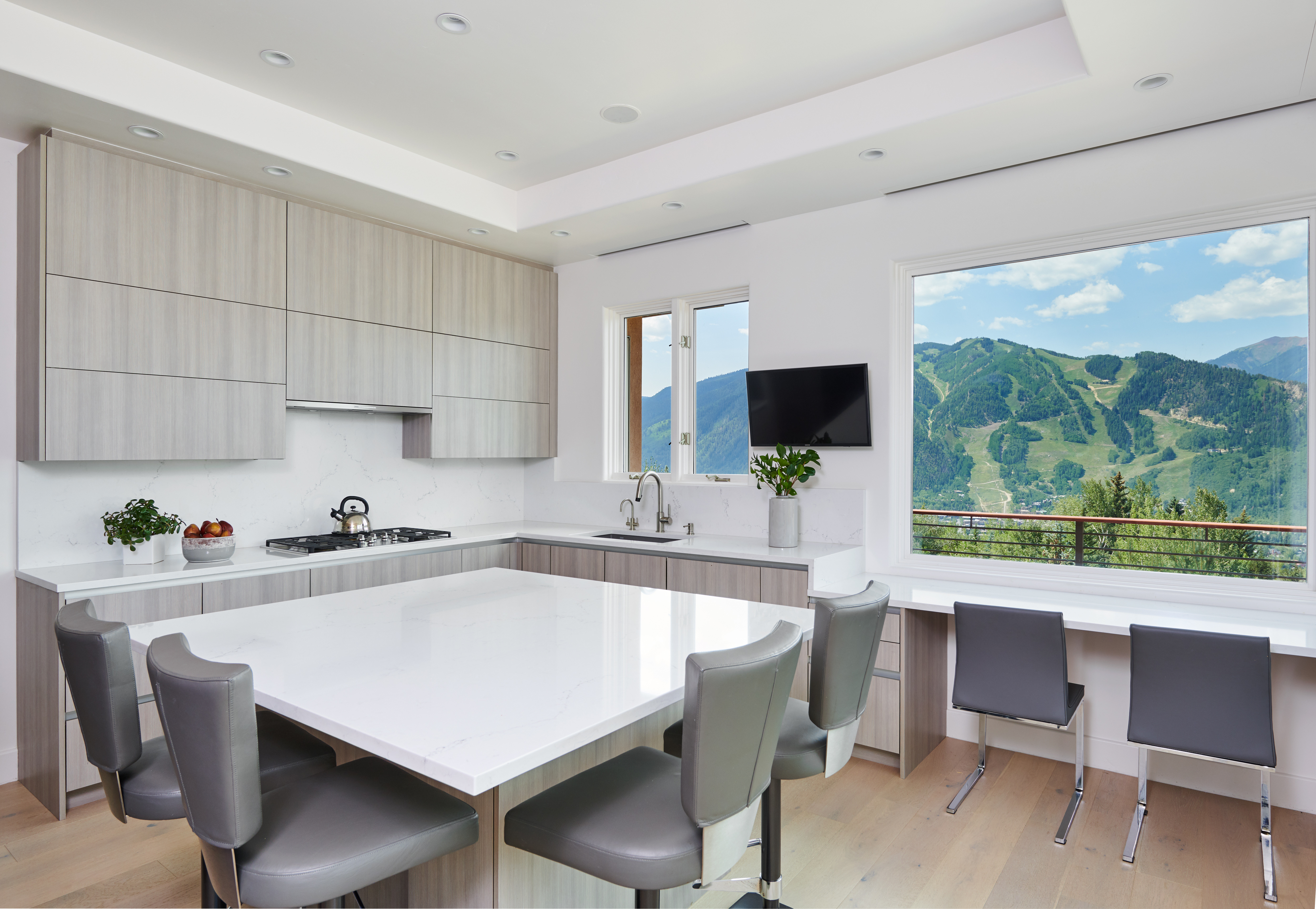 This sleek, modern kitchen features light wood cabinetry, a large white quartz island, and high-end stainless steel appliances. A large window provides a breathtaking, panoramic view of the surrounding mountains, seamlessly blending indoor luxury with the natural landscape. The space is bright and airy, with a minimalist aesthetic and a dedicated breakfast bar area overlooking the scenery.