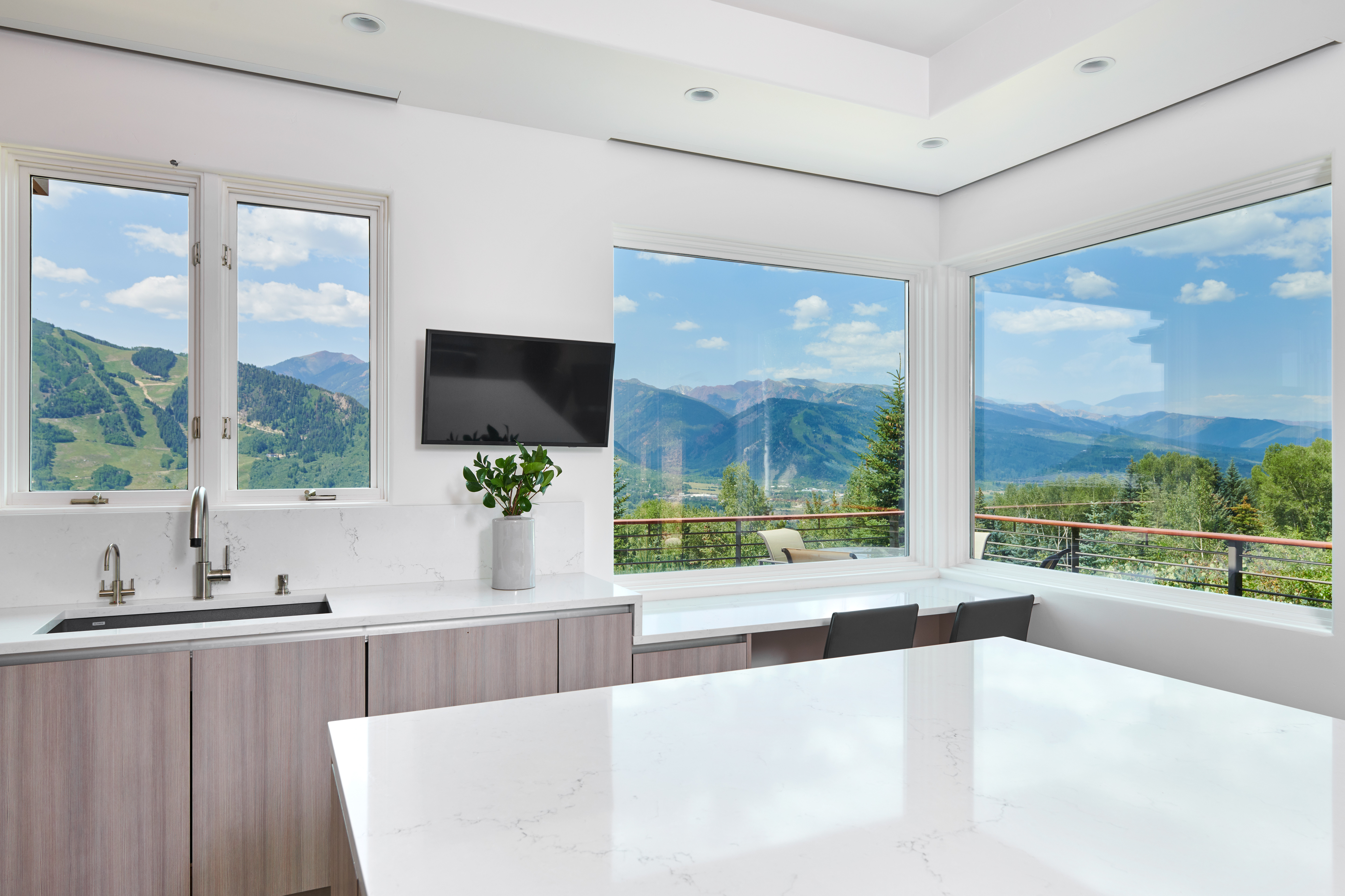 This modern kitchen features sleek, light-wood cabinetry paired with pristine white quartz countertops and a minimalist sink area. A wall-mounted television and a potted plant add a touch of personality, while expansive corner windows frame a breathtaking, panoramic view of lush green mountains. The bright, airy atmosphere is enhanced by recessed lighting and a clean, contemporary design aesthetic.