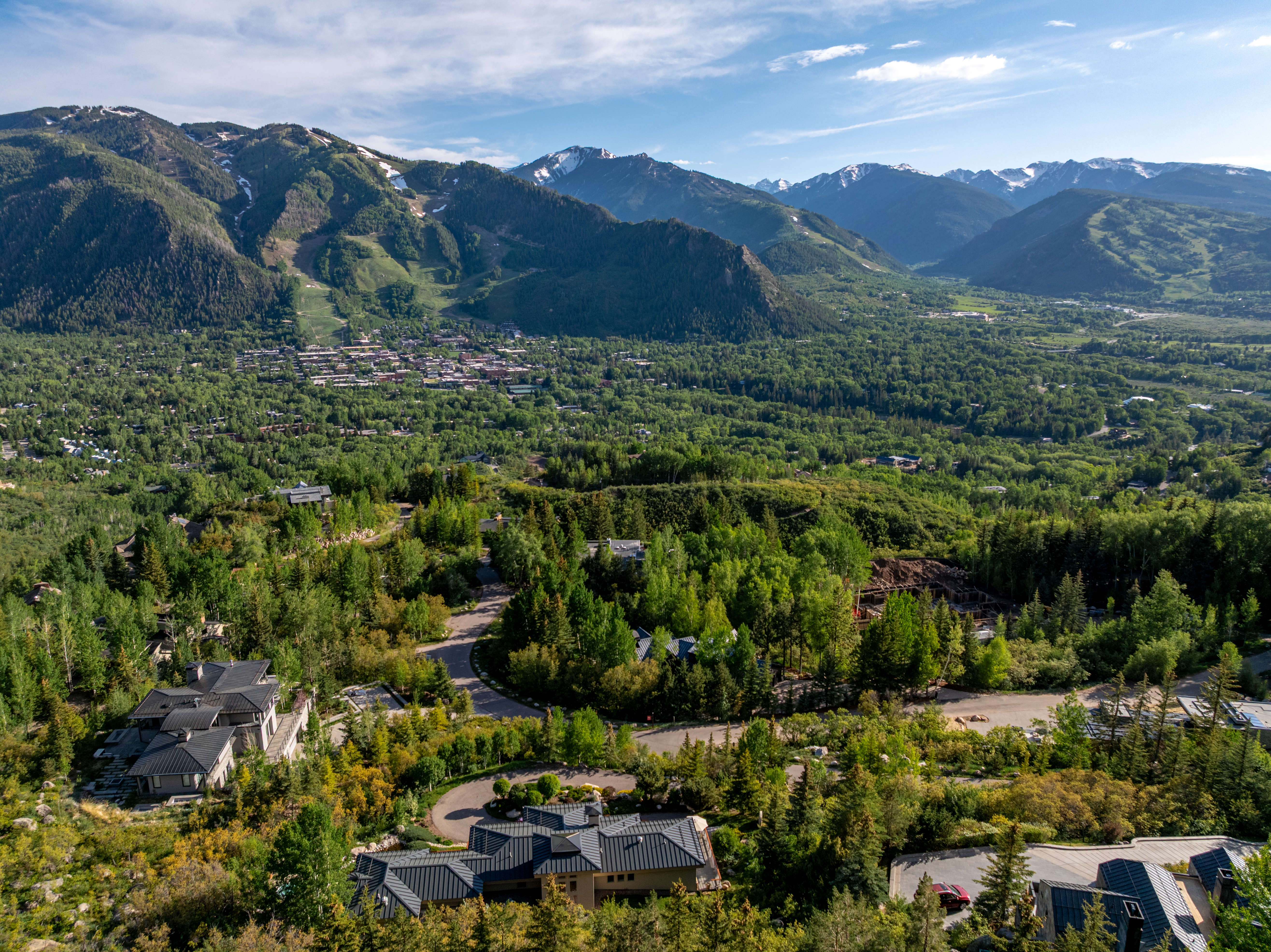 This high-angle aerial shot captures a luxurious mountain estate nestled within a lush, forested landscape overlooking a sprawling valley town. The property features multiple modern homes with dark metal roofs, winding driveways, and expansive views of the surrounding snow-capped peaks. The perspective emphasizes the serene, secluded nature of the location while highlighting its proximity to the vibrant town below.