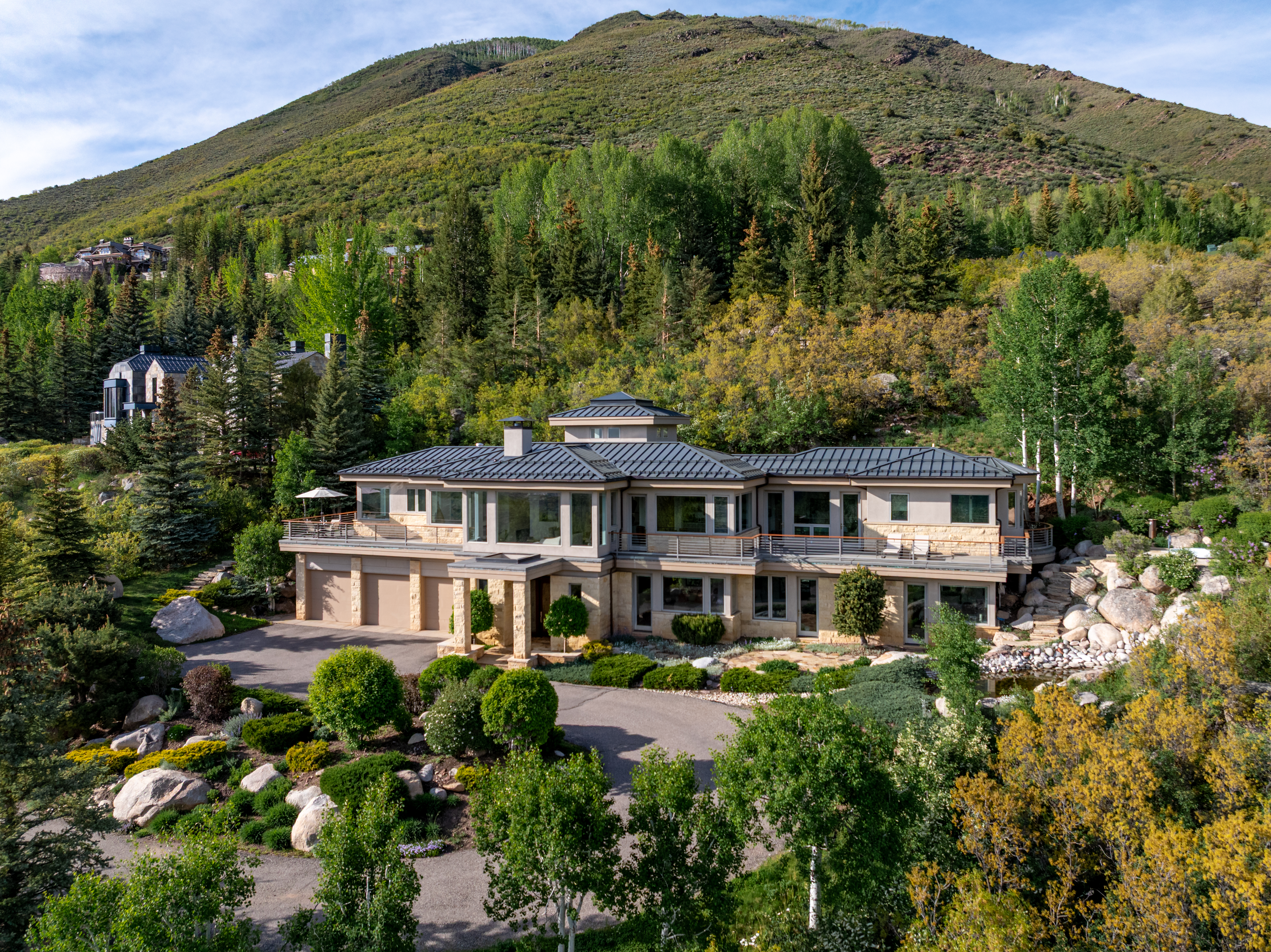 This stunning aerial view captures a luxurious mountain estate nestled into a lush, forested hillside. The home features a contemporary architectural design with a dark metal roof, stone accents, and expansive balconies that offer panoramic views of the surrounding landscape. A winding driveway leads to the multi-car garage, while the property is beautifully integrated into the natural terrain with mature trees and rock landscaping.
