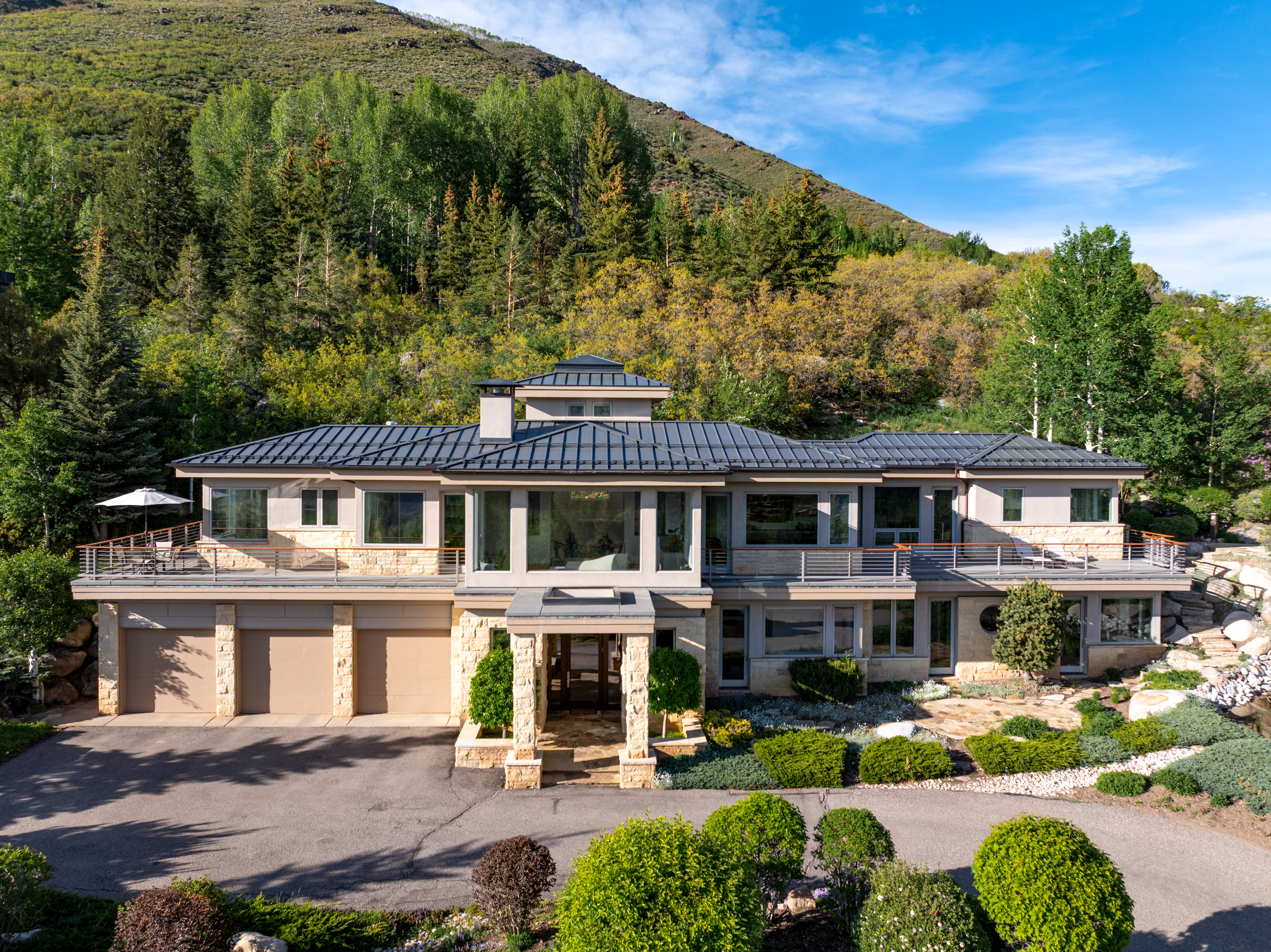 This stunning contemporary mountain estate features a multi-level design with a blend of natural stone and modern metal roofing. The home boasts expansive upper-level balconies, large floor-to-ceiling windows, and a three-car garage, all set against a lush, forested hillside backdrop. The cinematic perspective captures the grandeur of the architecture, emphasizing its seamless integration into the rugged, scenic landscape.