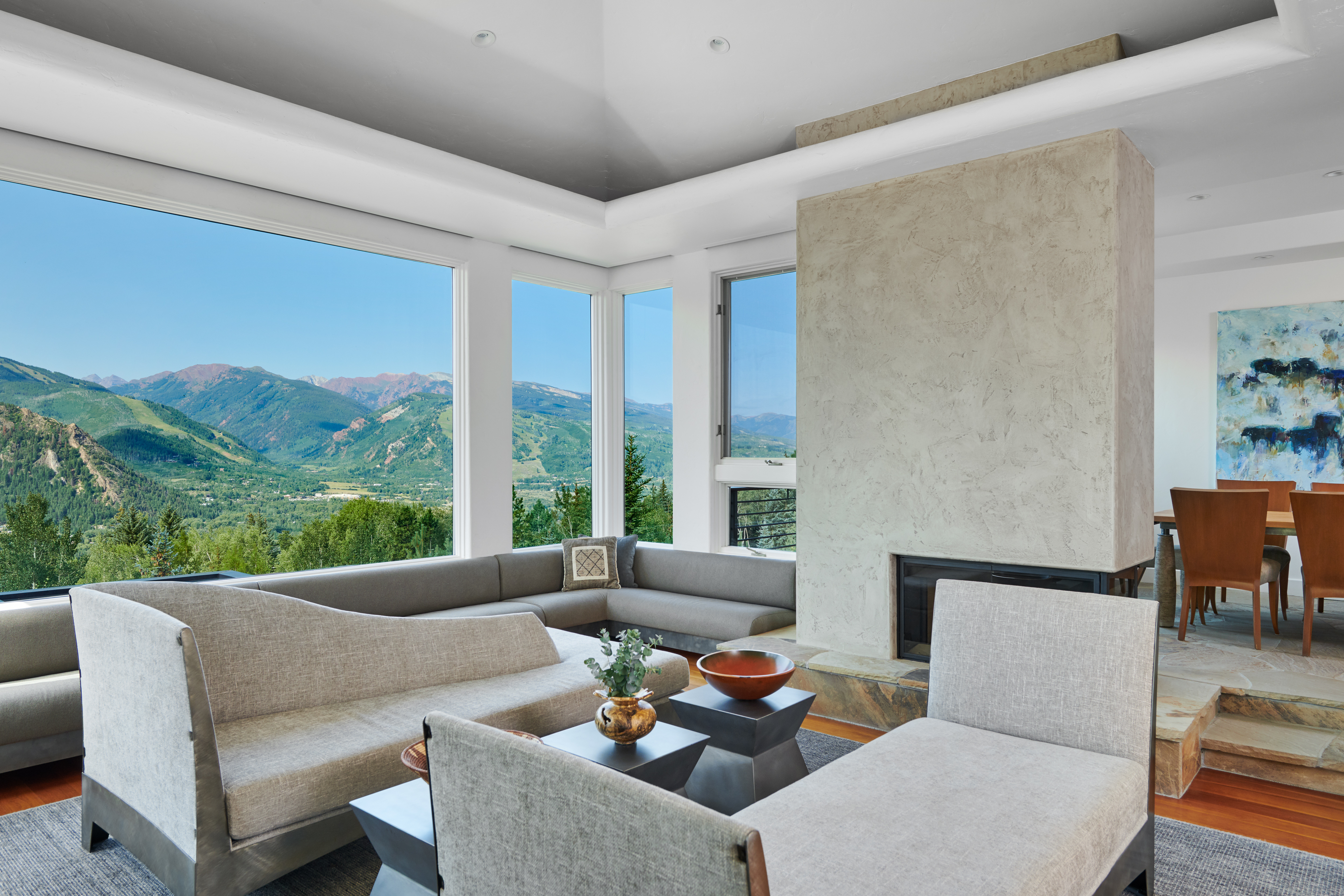 This sophisticated living room features a contemporary open-concept design with a large sectional sofa and a central fireplace finished in textured stone. Expansive floor-to-ceiling windows frame a breathtaking panoramic view of the surrounding mountains and lush valley. The space is accented by modern furniture, a neutral color palette, and a step-up dining area, creating an atmosphere of refined luxury and comfort.