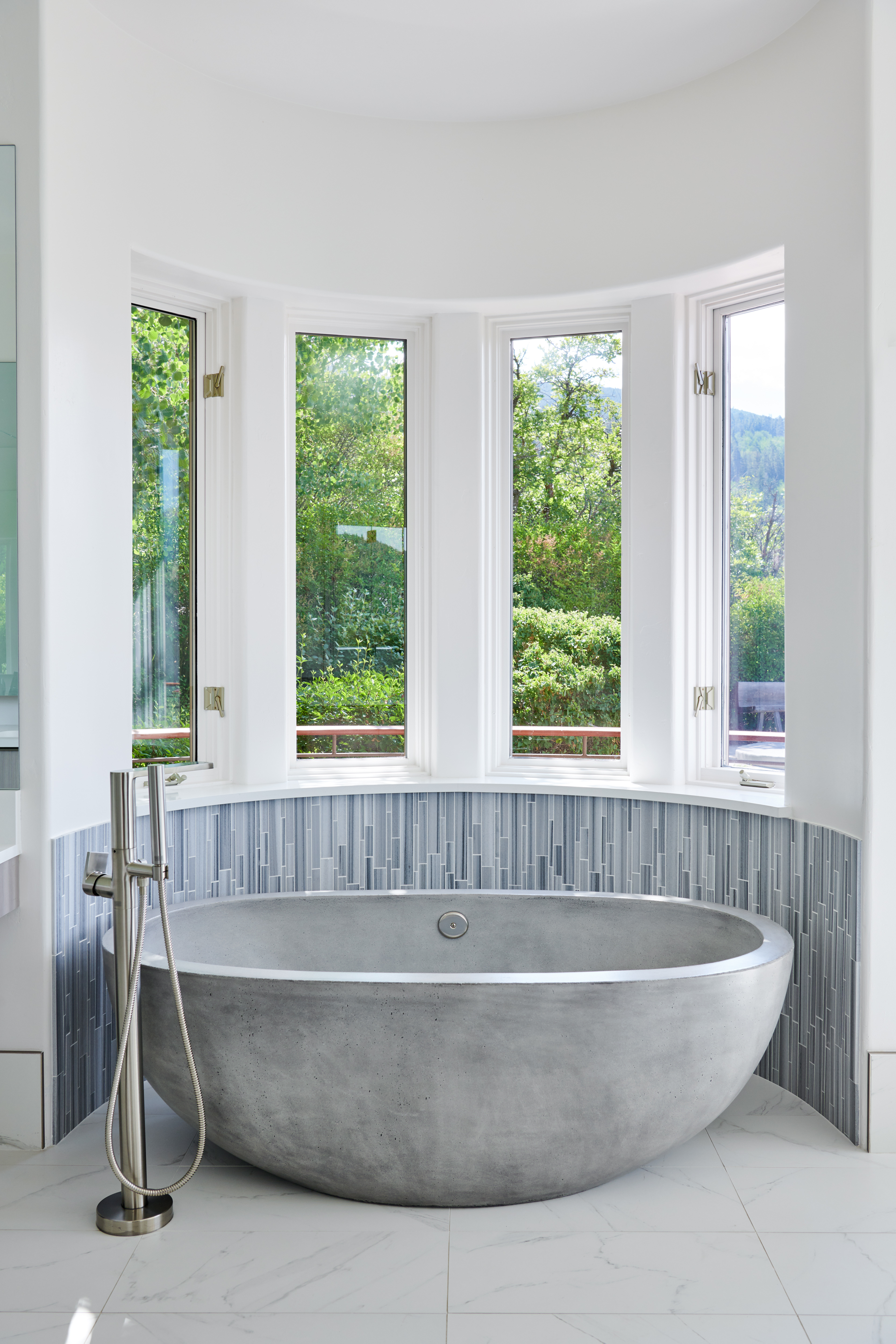 This luxurious primary bathroom features a striking, deep concrete soaking tub positioned centrally in a curved bay window alcove. The space is accented by a textured, vertical blue-grey tile backsplash and sleek, modern brushed-nickel plumbing fixtures. The bright, white-walled room is grounded by polished white marble-look flooring, creating a serene and spa-like atmosphere.