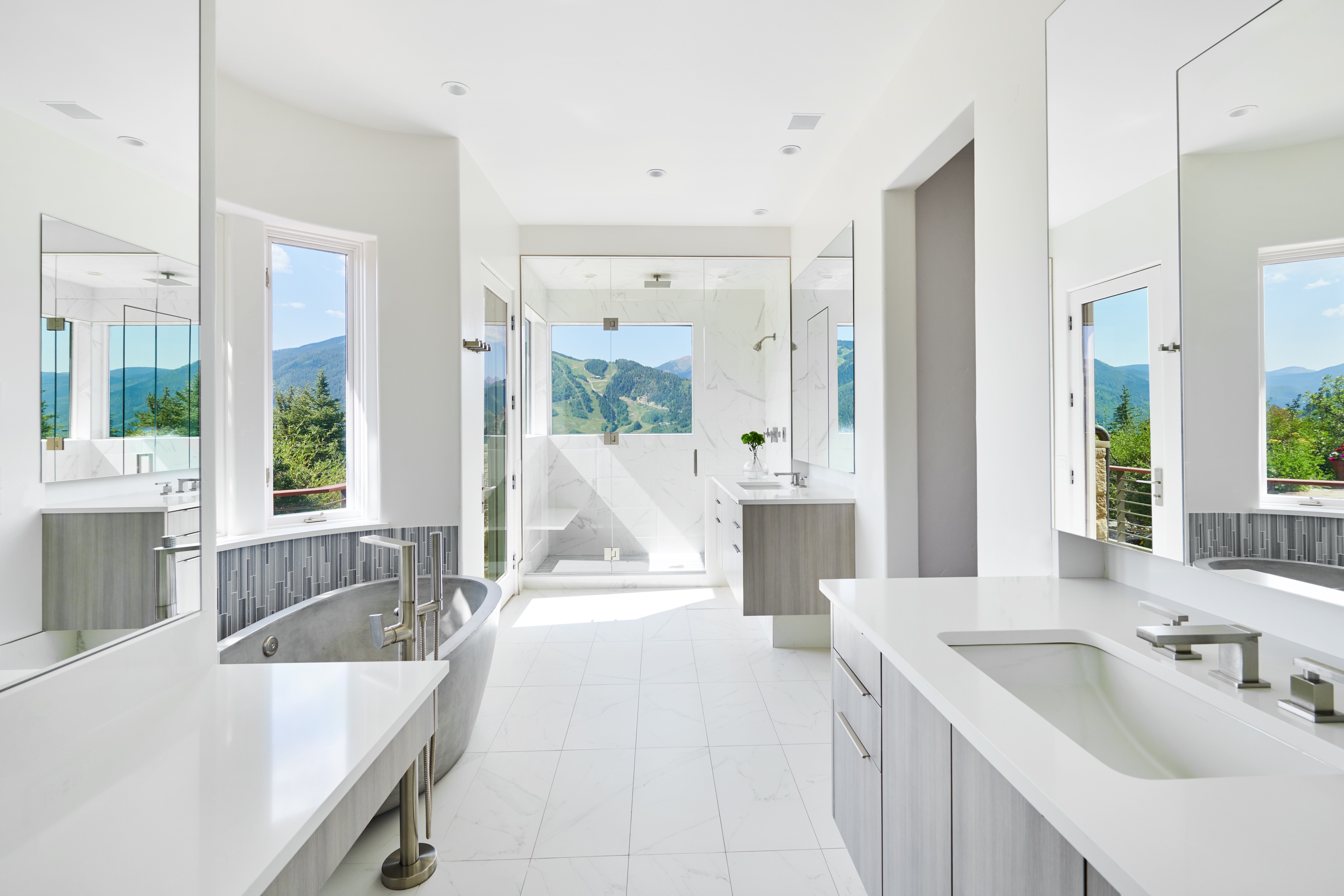 This luxurious primary bathroom features a sleek, modern design with a freestanding stone soaking tub, dual floating vanities, and a spacious glass-enclosed walk-in shower. Large windows frame breathtaking mountain views, bathing the white marble-tiled space in natural light. The clean lines and high-end finishes create a serene, spa-like atmosphere perfect for a high-end mountain retreat.
