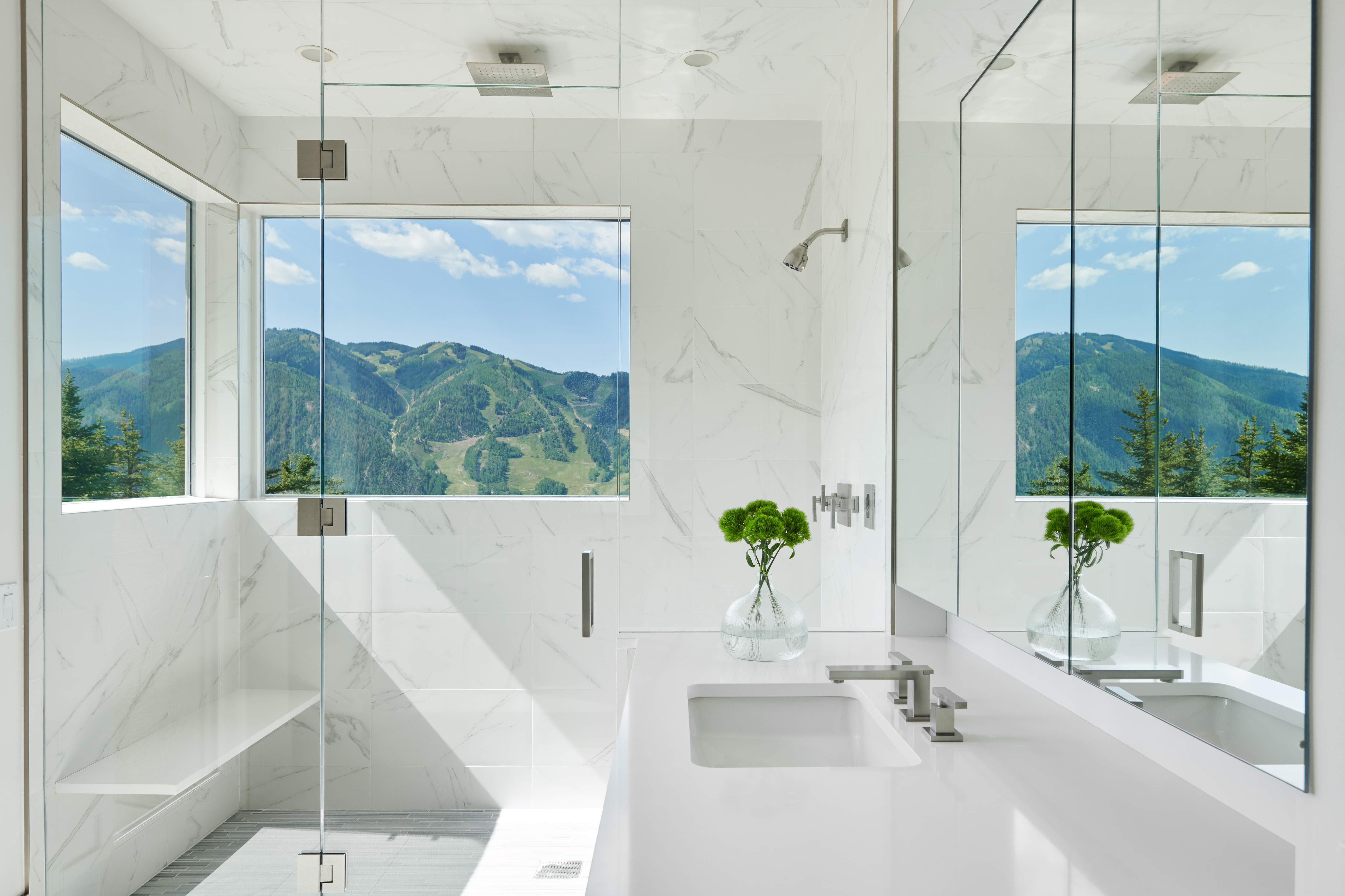 This luxurious primary bathroom features a sleek, modern design with floor-to-ceiling white marble tiling and a seamless glass-enclosed shower. A large window provides a breathtaking, unobstructed view of lush green mountains, creating a serene and spa-like atmosphere. The minimalist vanity with a white countertop and a clear glass vase with greenery adds a touch of elegance to the bright, airy space.