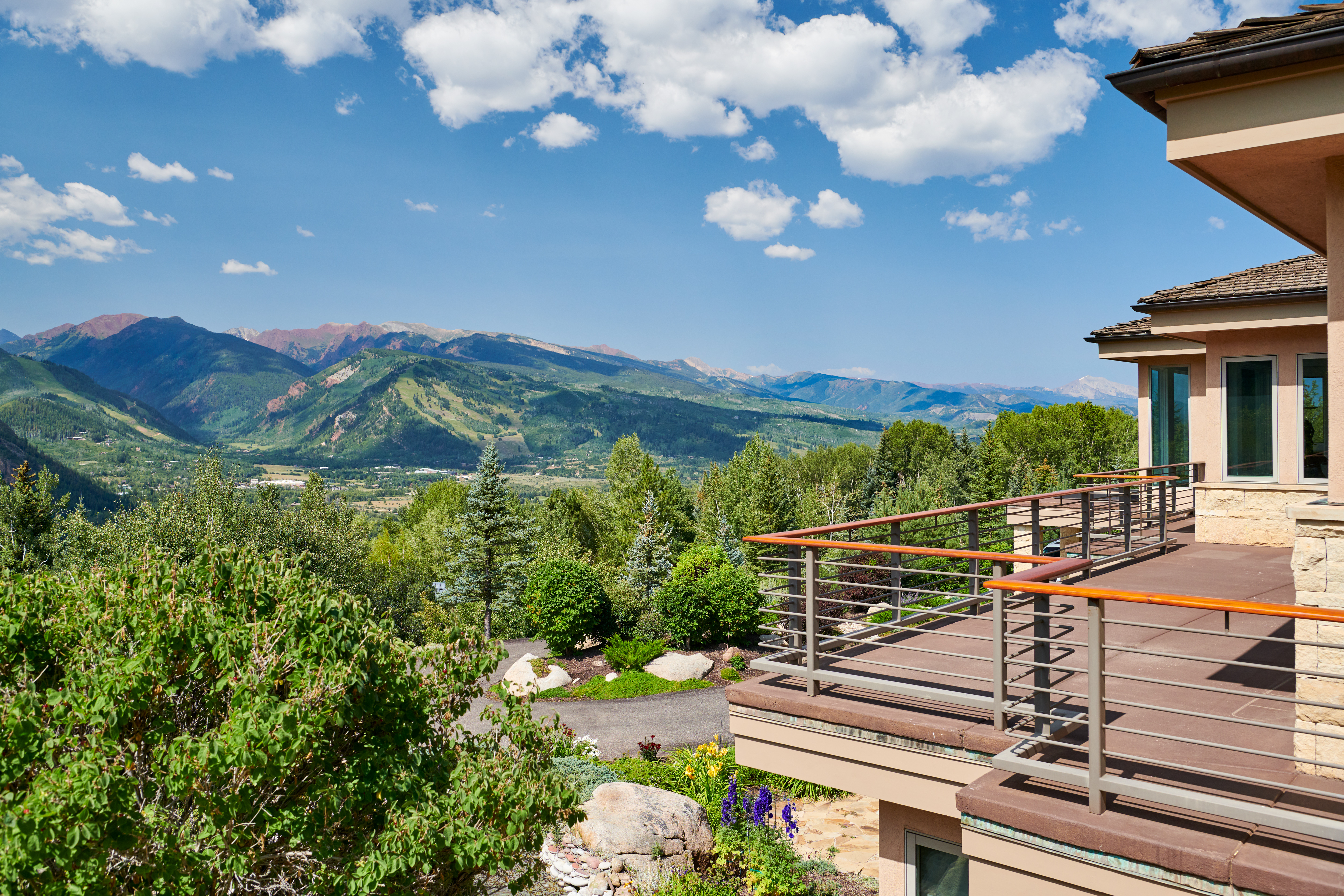 This high-angle shot captures a spacious, multi-level deck featuring modern metal railings with warm wood handrails, overlooking a breathtaking panoramic view of lush green valleys and majestic, snow-dusted mountain peaks. The scene is bathed in bright, natural sunlight under a clear blue sky, emphasizing the home's seamless integration with its stunning alpine surroundings. The foreground includes vibrant landscaping and natural rock elements, creating a serene and luxurious outdoor living space.