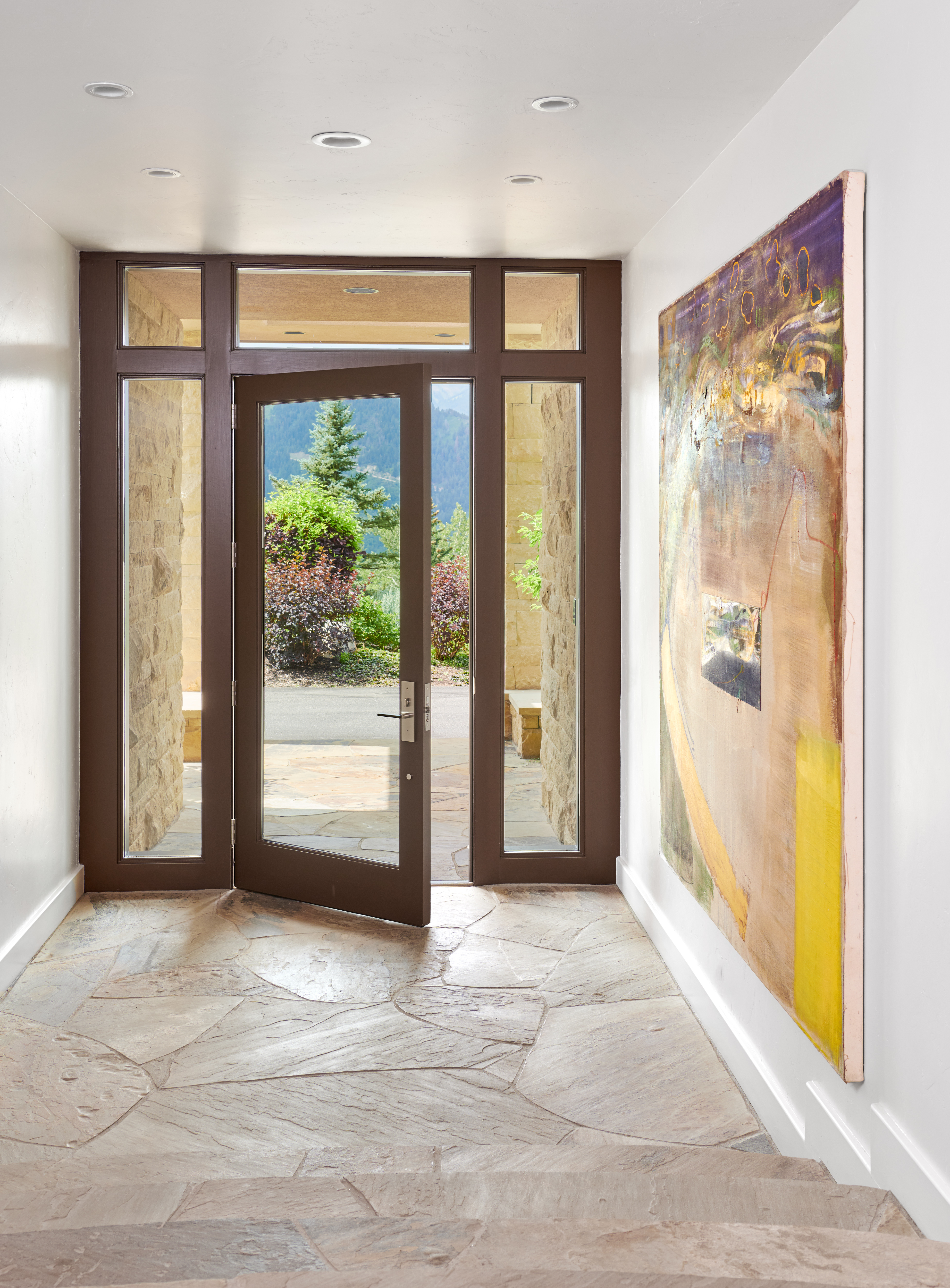 This inviting entryway features a grand, open-door design that frames a scenic mountain view, blending indoor and outdoor living. The space is characterized by natural stone flooring, clean white walls, and a large, vibrant abstract painting that serves as a focal point. The perspective is from the bottom of a short staircase, looking up toward the front door, creating a sense of arrival and architectural elegance.