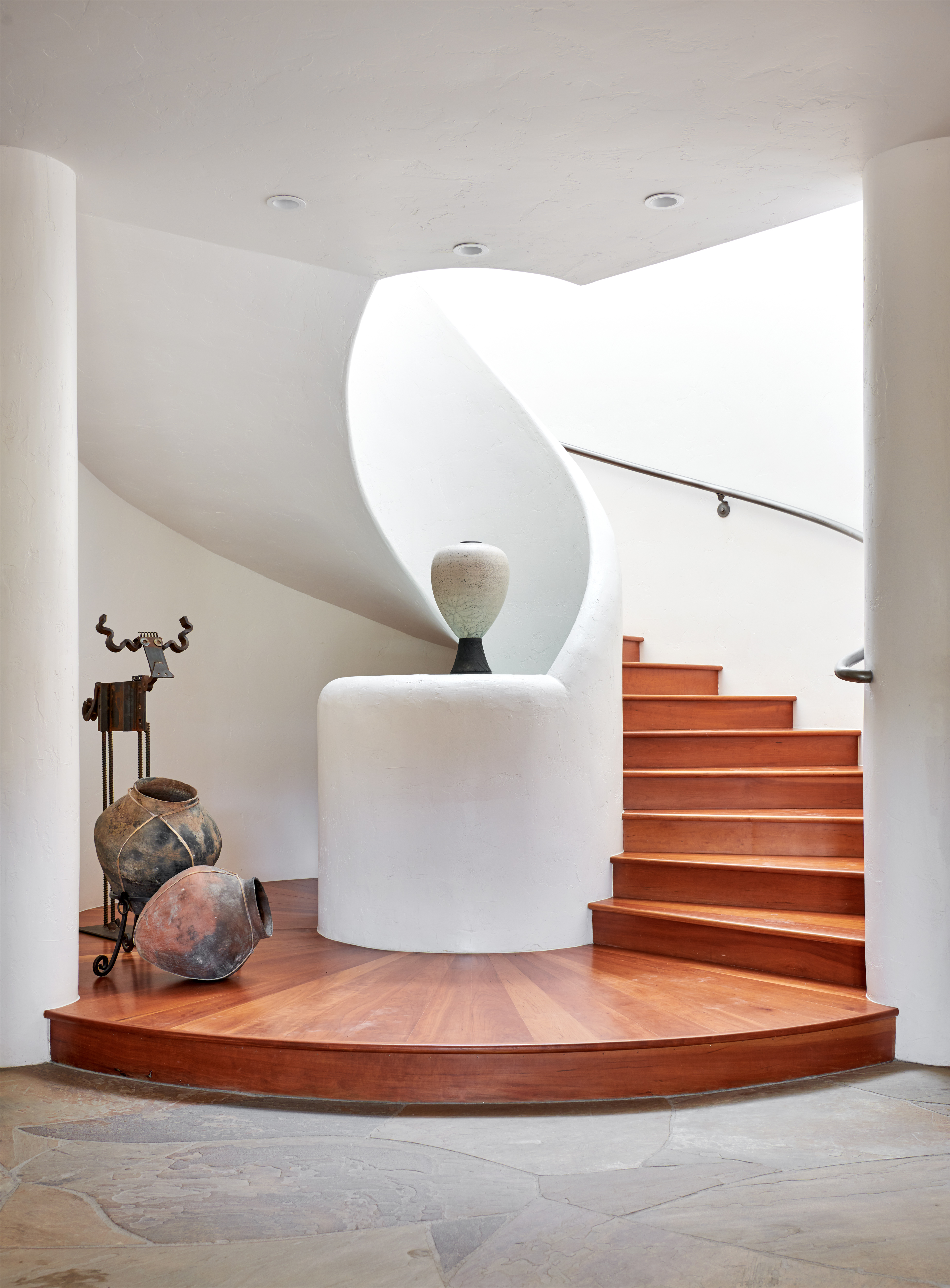 This elegant entryway features a striking, sculptural spiral staircase with warm wooden treads and a smooth, white curved wall. The base of the stairs is anchored by a circular wooden landing, complemented by rustic pottery and an artistic metal sculpture. The overall aesthetic is a sophisticated blend of organic, modern, and Southwestern design elements.