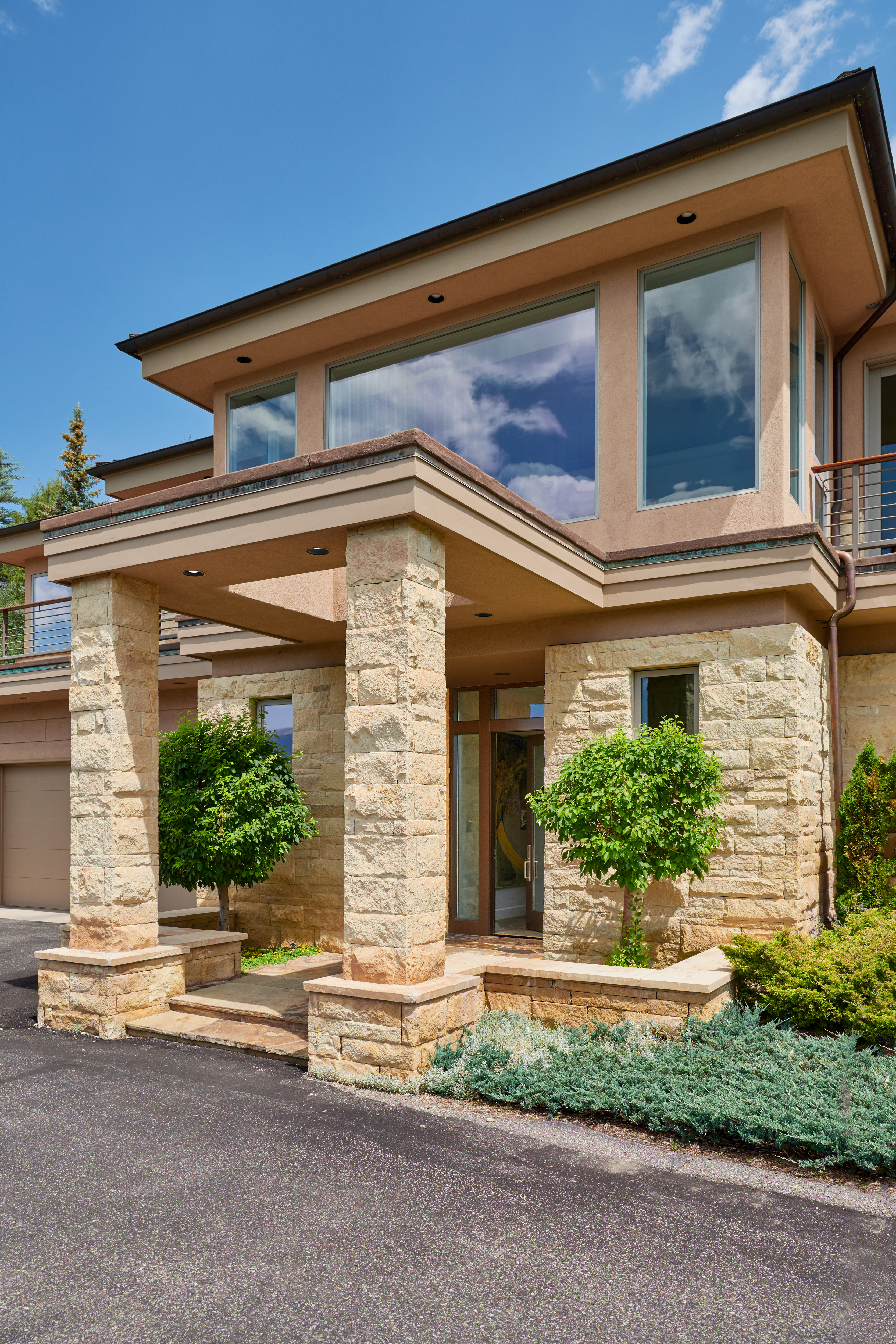 This striking entryway features a prominent stone-clad portico supported by two massive, textured stone pillars, creating a grand and welcoming focal point. The home showcases a contemporary architectural style with warm-toned stucco walls, large expansive windows reflecting the sky, and manicured landscaping that softens the stone elements. The perspective is a low-angle, eye-level shot that emphasizes the height and solid construction of the home's entrance.