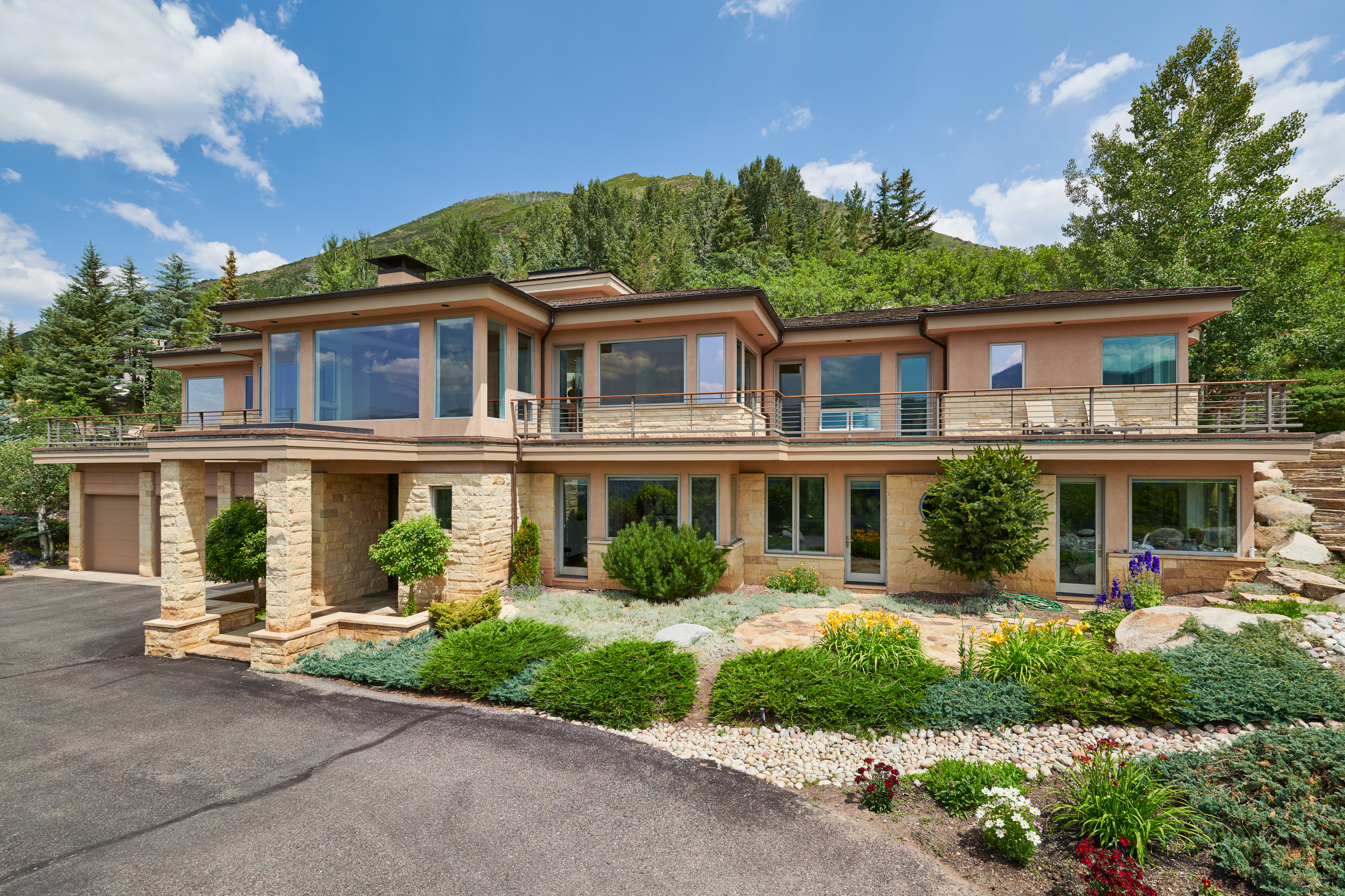 This stunning multi-level mountain residence features a sophisticated blend of natural stone masonry and warm-toned stucco, perfectly integrated into a lush, landscaped hillside. The home boasts expansive floor-to-ceiling windows and a wrap-around balcony that offers panoramic views of the surrounding forest and peaks. A paved driveway leads to the garage, while the foreground showcases a meticulously maintained garden with vibrant perennials and ornamental shrubs.