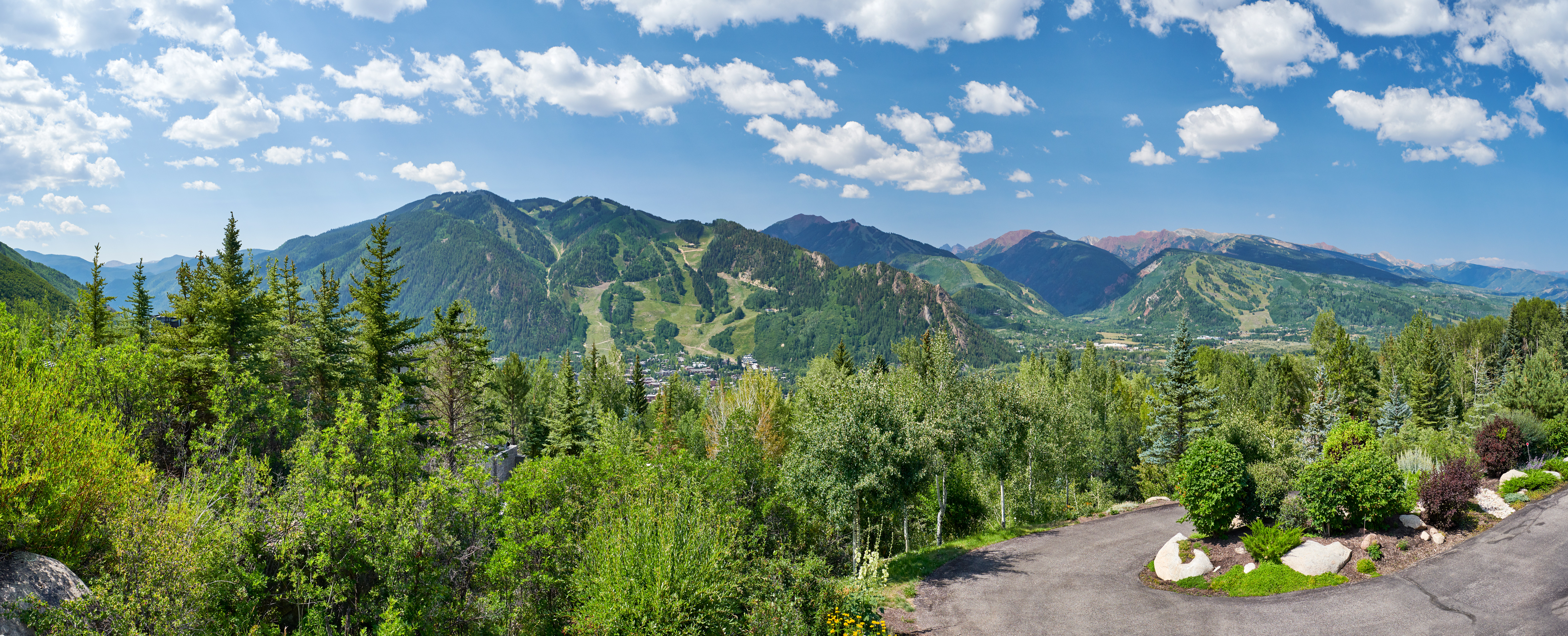 This panoramic view captures a breathtaking mountain landscape, likely taken from a high-elevation residential property. The foreground features lush green trees and a paved driveway, leading the eye toward a sprawling valley nestled between majestic, sun-drenched mountain peaks under a bright blue sky with scattered clouds. The perspective is cinematic and expansive, emphasizing the serene, natural beauty and the prime location of the property.