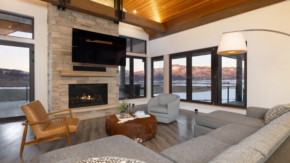 This living room features a striking stone fireplace with a mounted television and a natural light-filled atmosphere provided by expansive windows that frame mountain views. The interior design blends rustic warmth with modern comfort, showcased by wooden ceiling beams, a leather armchair, a rounded hide-covered coffee table, and a large grey sectional. The open-plan layout creates a welcoming, sophisticated space perfect for relaxation, highlighting the connection between the high-end interior architecture and the scenic outdoor surroundings.