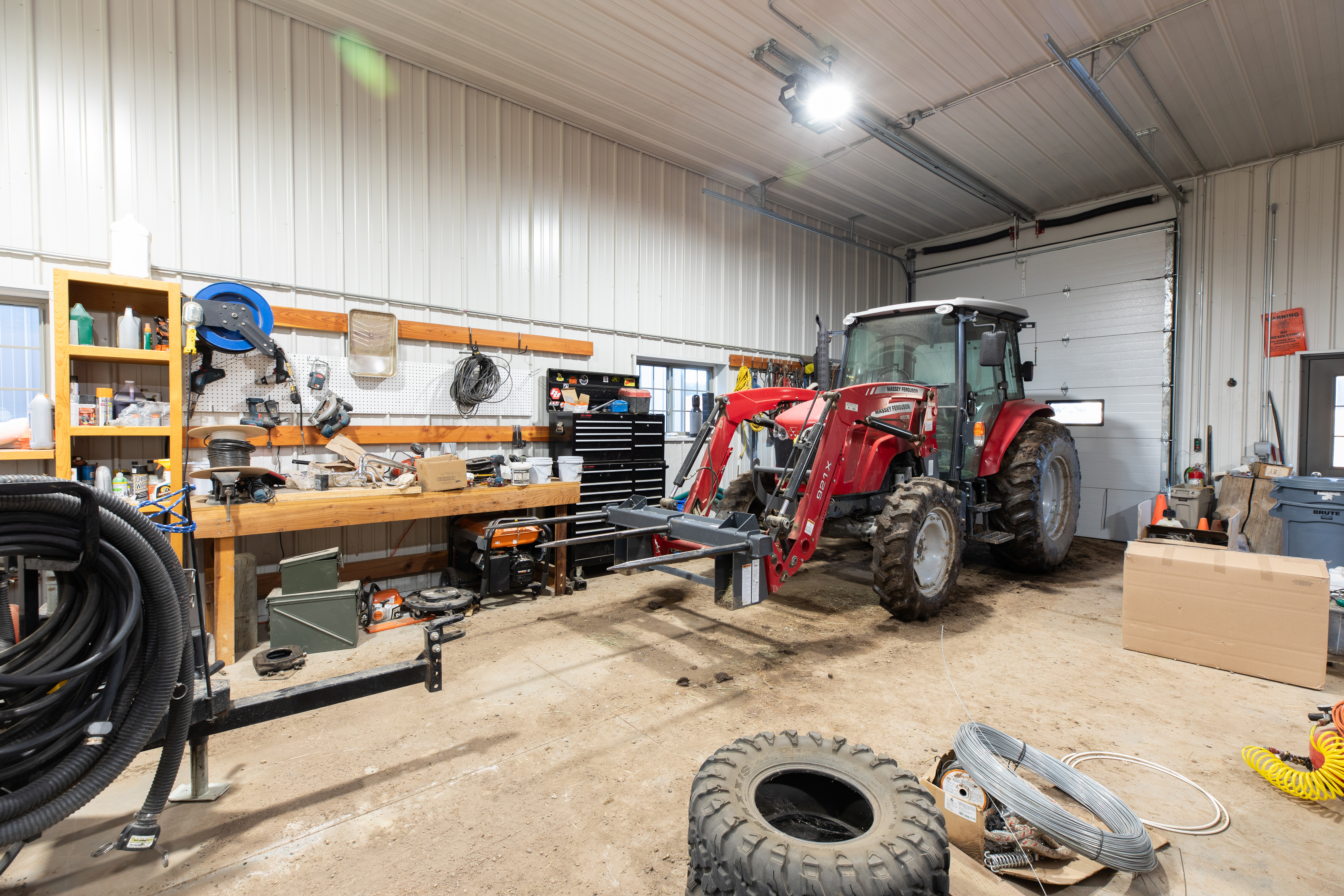 This interior view of a spacious, high-ceiling workshop or garage features corrugated metal walls and concrete flooring, perfect for heavy-duty projects or equipment storage. The space is anchored by a large red tractor with a front-loader attachment, complemented by a well-organized workbench with shelves and tool storage. A large overhead rolling door and bright overhead lighting make this a highly functional and practical workspace.