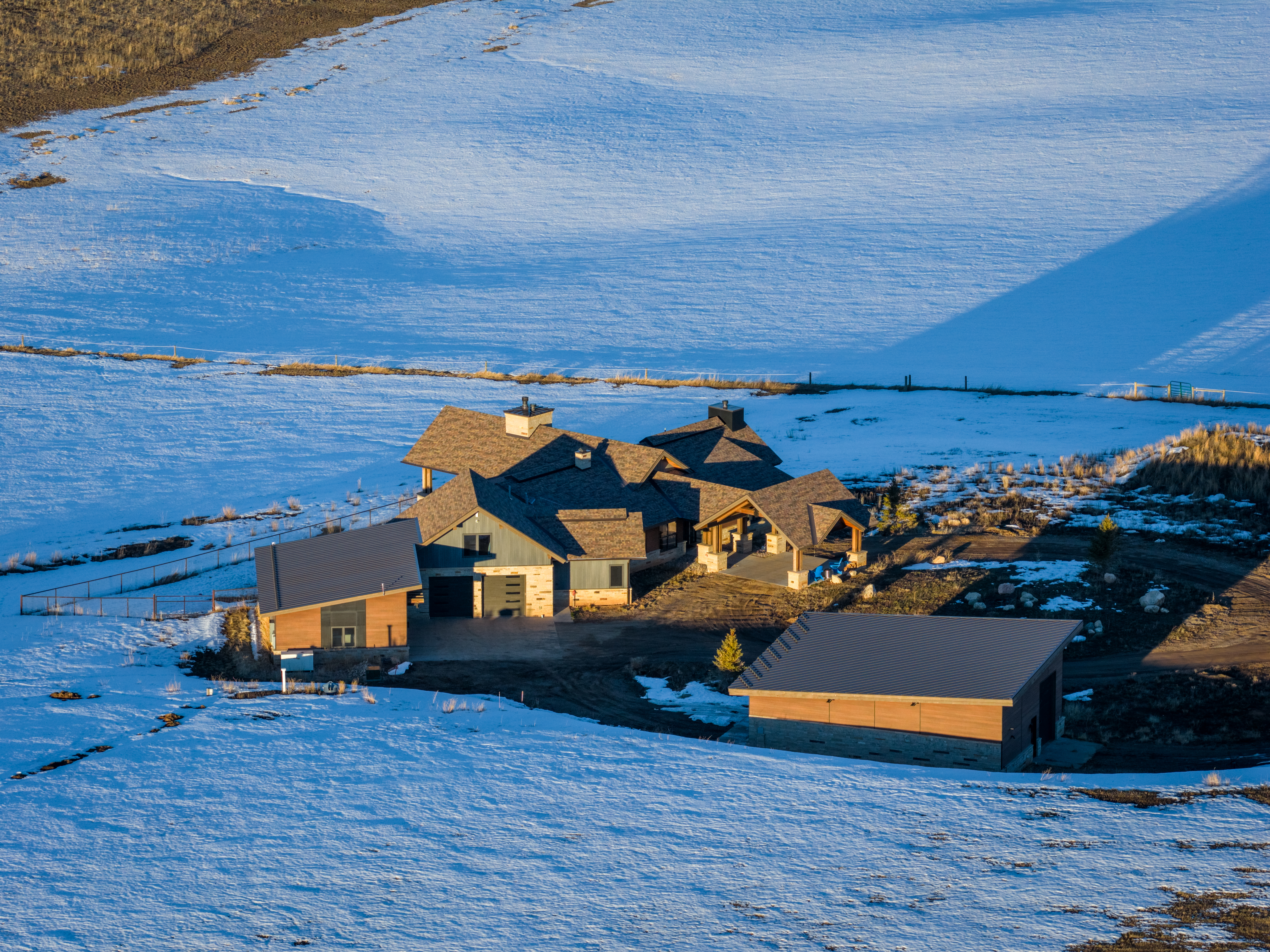 This is an impressive aerial view of a large, modern luxury mountain home situated in a sprawling, snow-covered rural landscape. The property features a complex roofline, stone and wood siding, and includes a detached garage or outbuilding, reflecting a high-end rustic architectural style. The scene suggests a remote, private retreat with expansive grounds during the early or late sunlight hours.