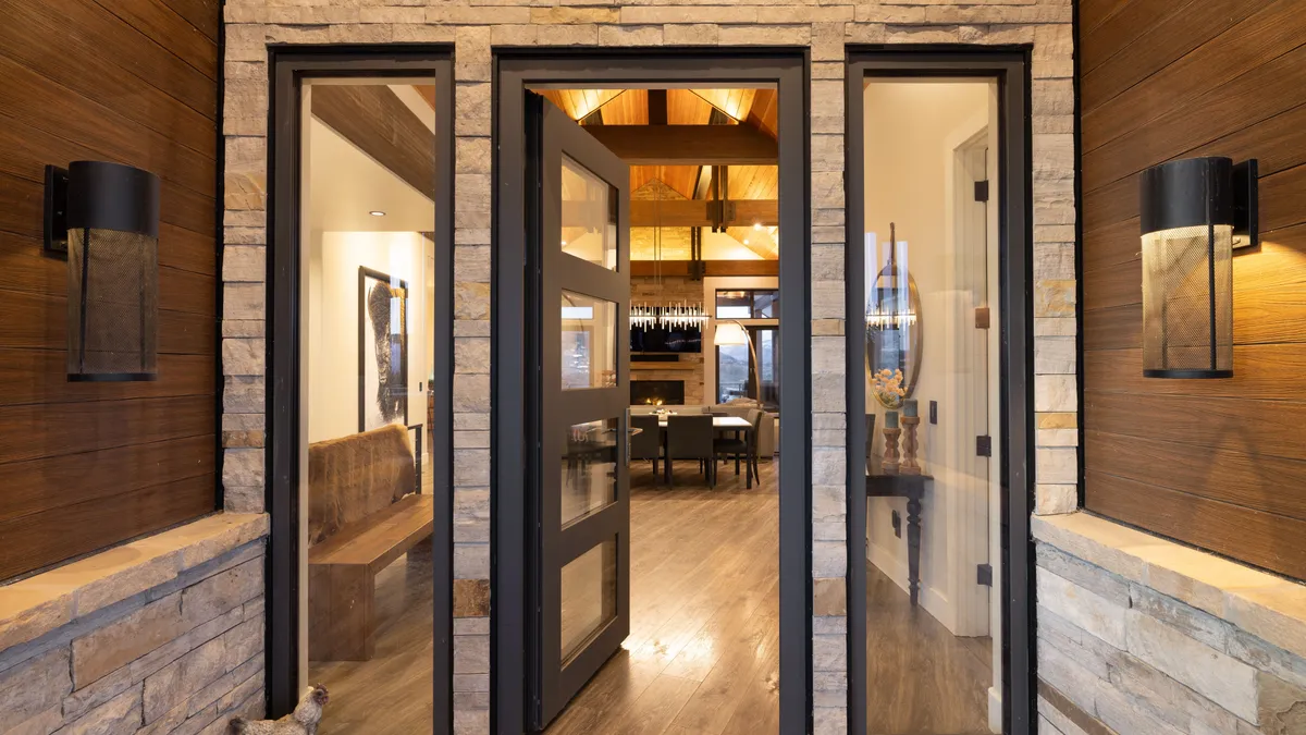 This stunning entryway features a modern, oversized front door flanked by two full-length sidelight windows, framed by textured limestone siding. The exterior design incorporates warm wood-paneled walls and sleek, cylindrical black mesh sconces that provide a sophisticated, contemporary rustic aesthetic. Through the glass, a glimpse into the open-concept interior reveals high vaulted ceilings, elegant lighting, and a view through to the dining and living spaces.