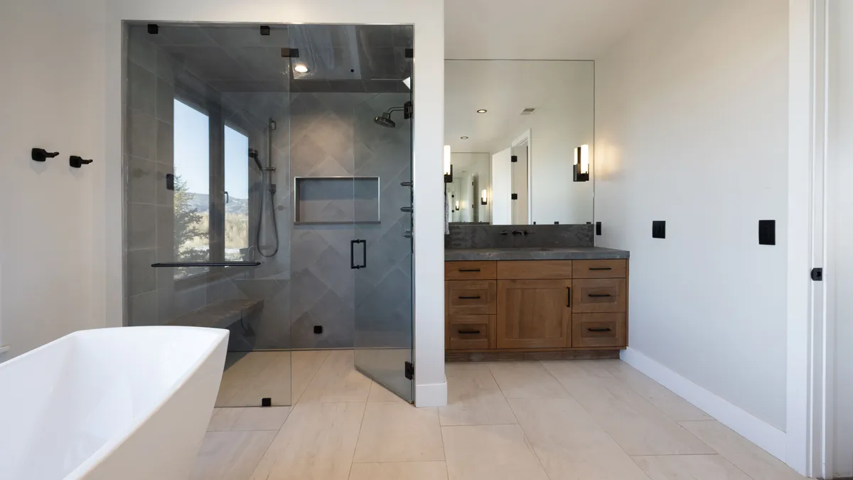This modern bathroom features a sleek glass-enclosed shower with gray tiled walls and a natural wood vanity topped with a dark stone countertop. A freestanding white soaking tub sits partially in view, creating a spa-like atmosphere. The combination of clean lines, muted tones, and contemporary light fixtures gives the space an elegant and sophisticated look.