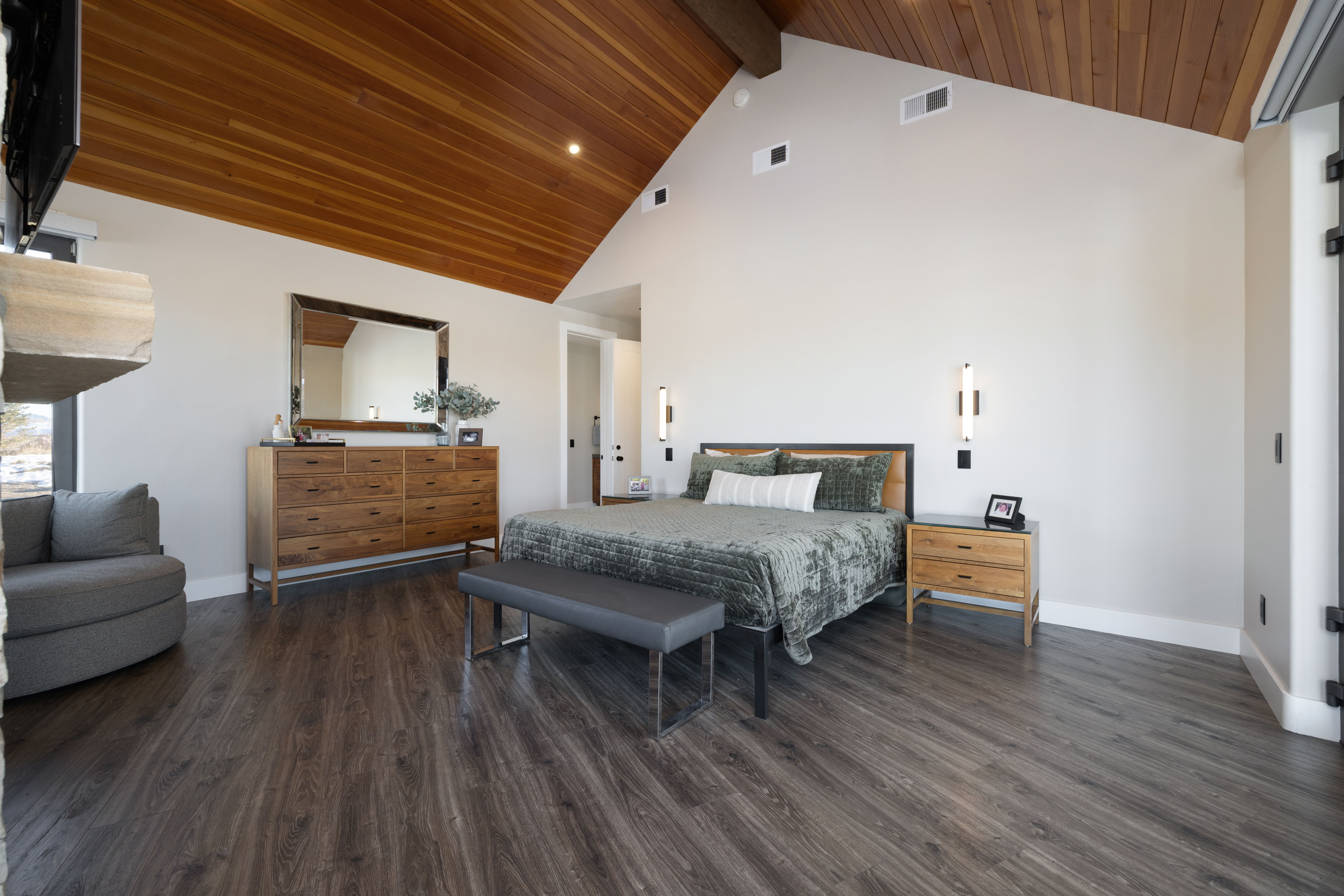 This spacious primary bedroom features a striking vaulted ceiling with warm wood tongue-and-groove paneling and a modern, airy design. The room is anchored by a comfortable bed accented with a velvet-textured green coverlet, flanked by contemporary nightstands and sleek wall-mounted lighting. With its large wooden dresser, gray accent chair corner, and clean, neutral walls, the space offers a sophisticated blend of rustic charm and modern luxury.
