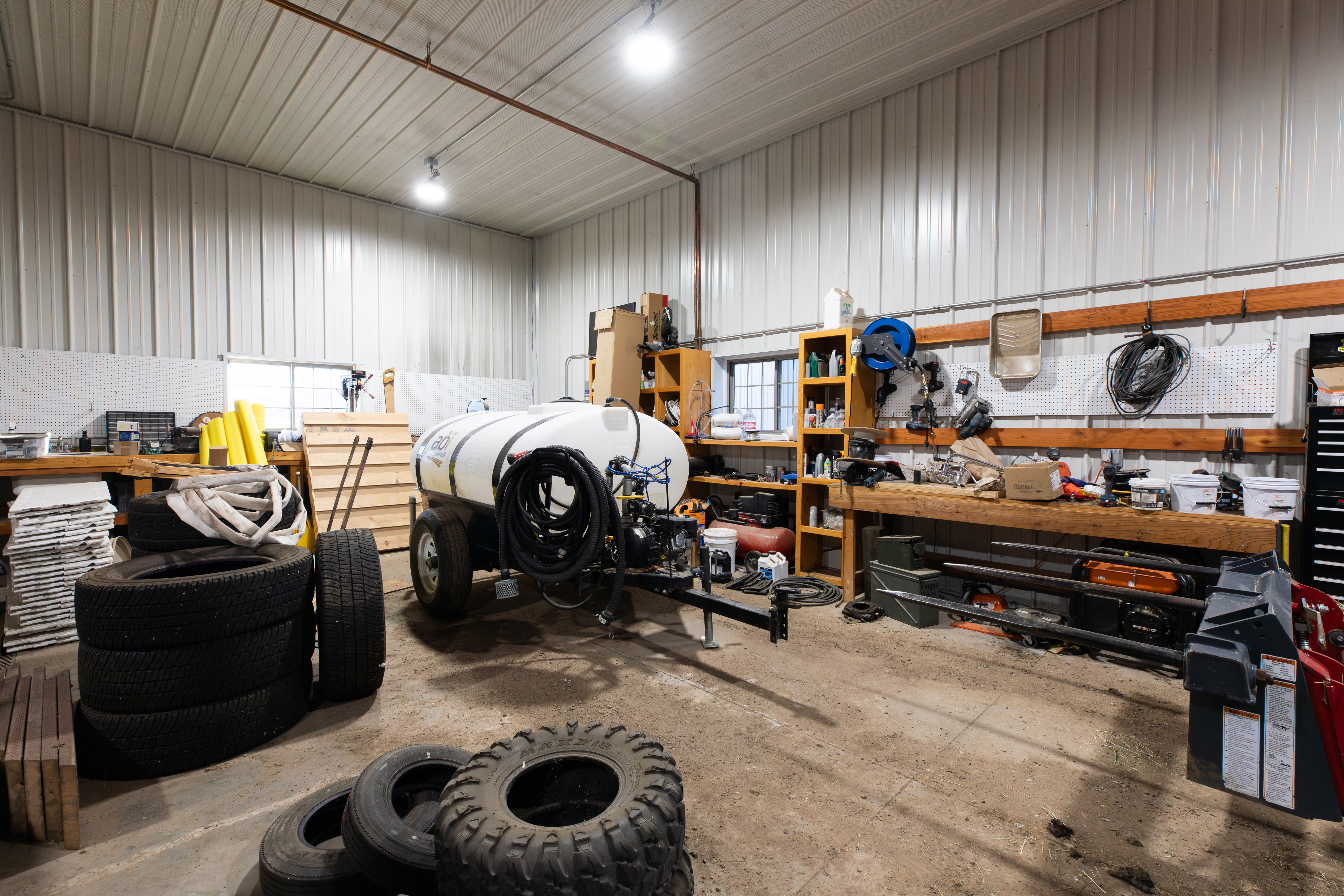 This interior view shows a spacious, functional workshop or utility barn. It features metal wall siding, pegboard organizational systems for tools, and a large workbench, making it ideal for storage or DIY projects. The space provides practical utility with ample room for heavy equipment, tires, and a trailer.