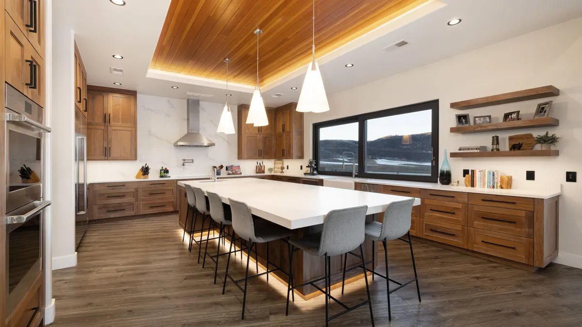 This stunning contemporary kitchen features warm wood cabinetry contrasted with crisp white countertops and a matching marble backsplash. The centerpiece is a large, island with integrated lighting and leather-upholstered bar stools, while a dramatic wood-plank ceiling adds texture and depth to the airy room. Large windows overlooking a scenic landscape provide ample natural light, perfectly balancing the warm organic tones with modern design elements.