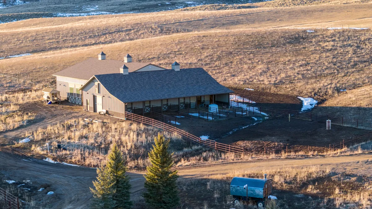 This elevated view shows a large, modern barn-style stable complex situated on a rural property. The structure features light-colored siding, a dark pitched roof with multiple cupolas, and integrated stalls connected to fenced exercise paddocks. The surrounding dry landscape and open terrain suggest a quiet, equestrian-focused rural retreat.