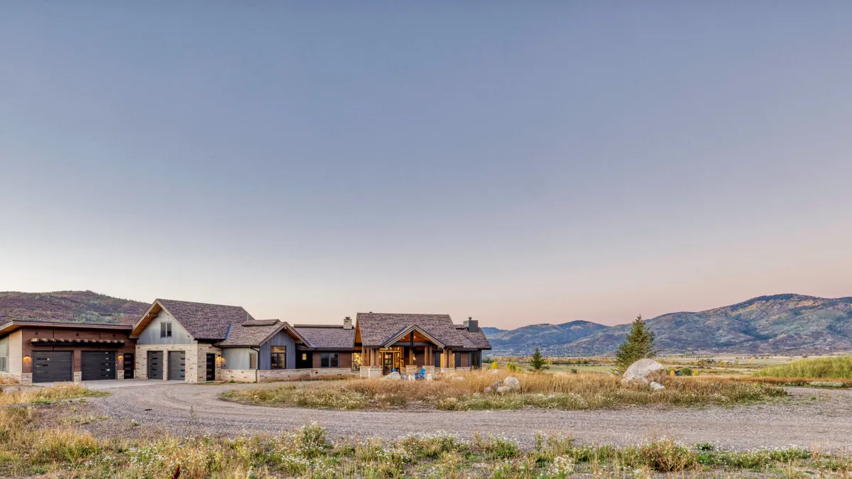 This stunning contemporary mountain estate features a harmonious blend of stone and dark metal siding, complemented by a multi-gabled roofline. The property includes an attached multi-car garage and a spacious outdoor entry area, all set against a breathtaking mountainous landscape. Its rustic yet modern architectural design offers a luxurious retreat feel, perfectly integrated into the sprawling natural terrain.