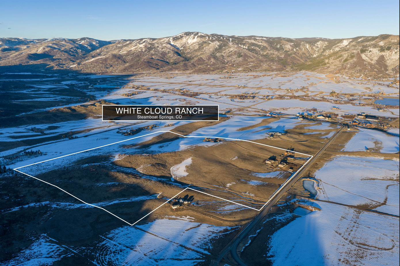 This is an aerial view of the White Cloud Ranch in Steamboat Springs, CO, highlighting a vast, expansive plot of land outlined in white. The property features rolling, snow-dusted terrain with existing rustic structures and direct access to nearby roadways. It offers a stunning, scenic perspective of the surrounding mountain landscape, emphasizing the rural, open character of the site.