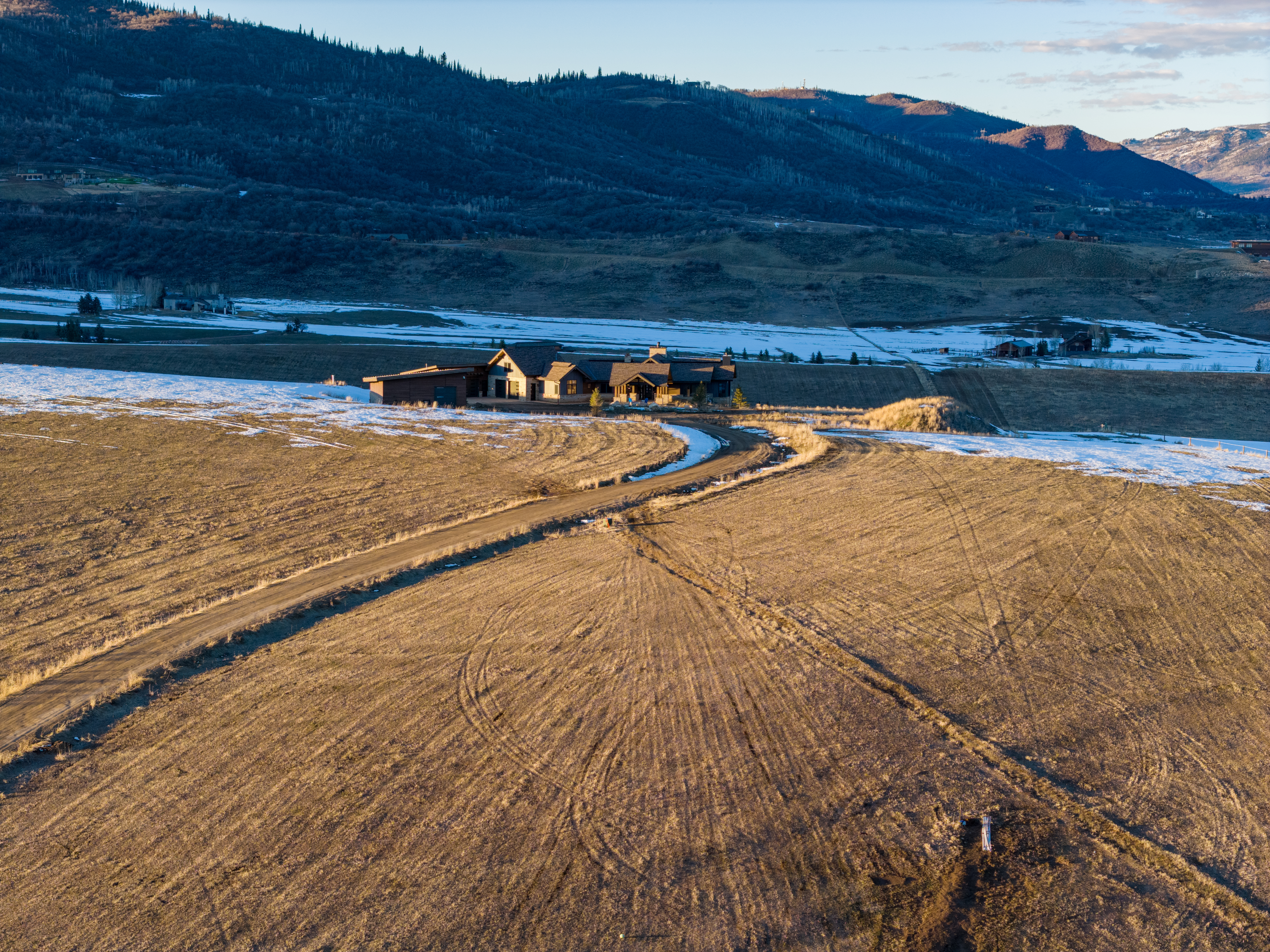This aerial view showcases a spacious, contemporary ranch-style home set amidst a vast, scenic rural landscape. The property features a long gravel driveway that curves through open fields, highlighting the expansive and private terrain surrounding the mountainous backdrop. The structure boasts high-quality materials and architectural design, perfectly integrated into the serene, natural environment.