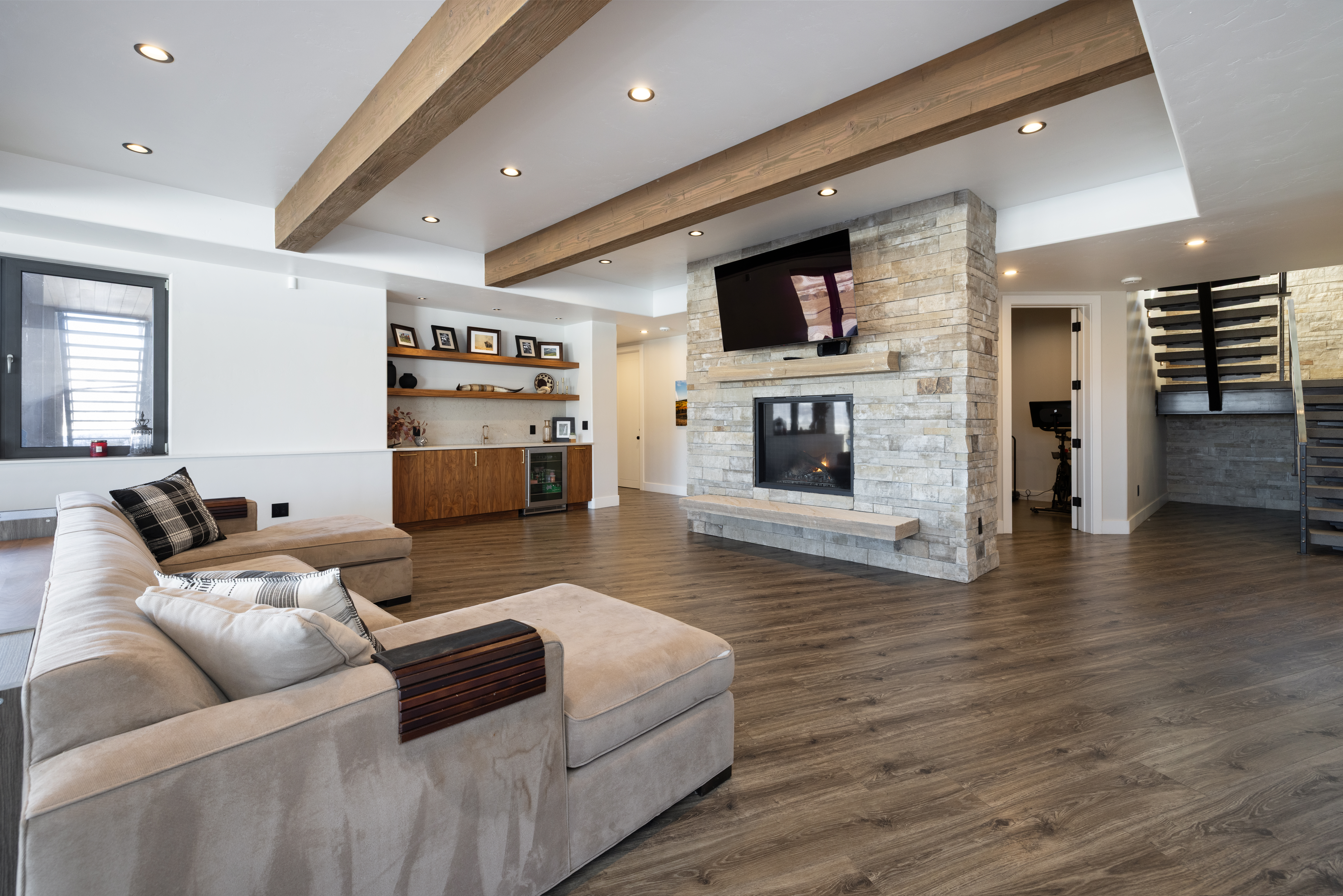 This spacious, open-concept living area features a modern rustic aesthetic with prominent wooden ceiling beams, a striking floor-to-ceiling stone fireplace, and light-toned, plush sectional seating. The space includes a convenient wet bar area with decorative shelving, wide-plank wood flooring, and a sleek metal staircase, creating a sophisticated and welcoming atmosphere for entertainment.