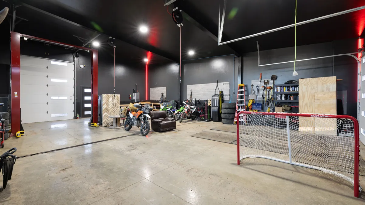 This spacious, modern garage features a high-ceiling design with sleek black walls and polished concrete floors. The area is well-equipped for projects and storage, including a built-in automotive lift, motorcycle parking, and organized wall-mounted tool racks. A Bauer hockey net and occasional furniture add to the functional, multi-purpose appeal of this expansive workshop space.